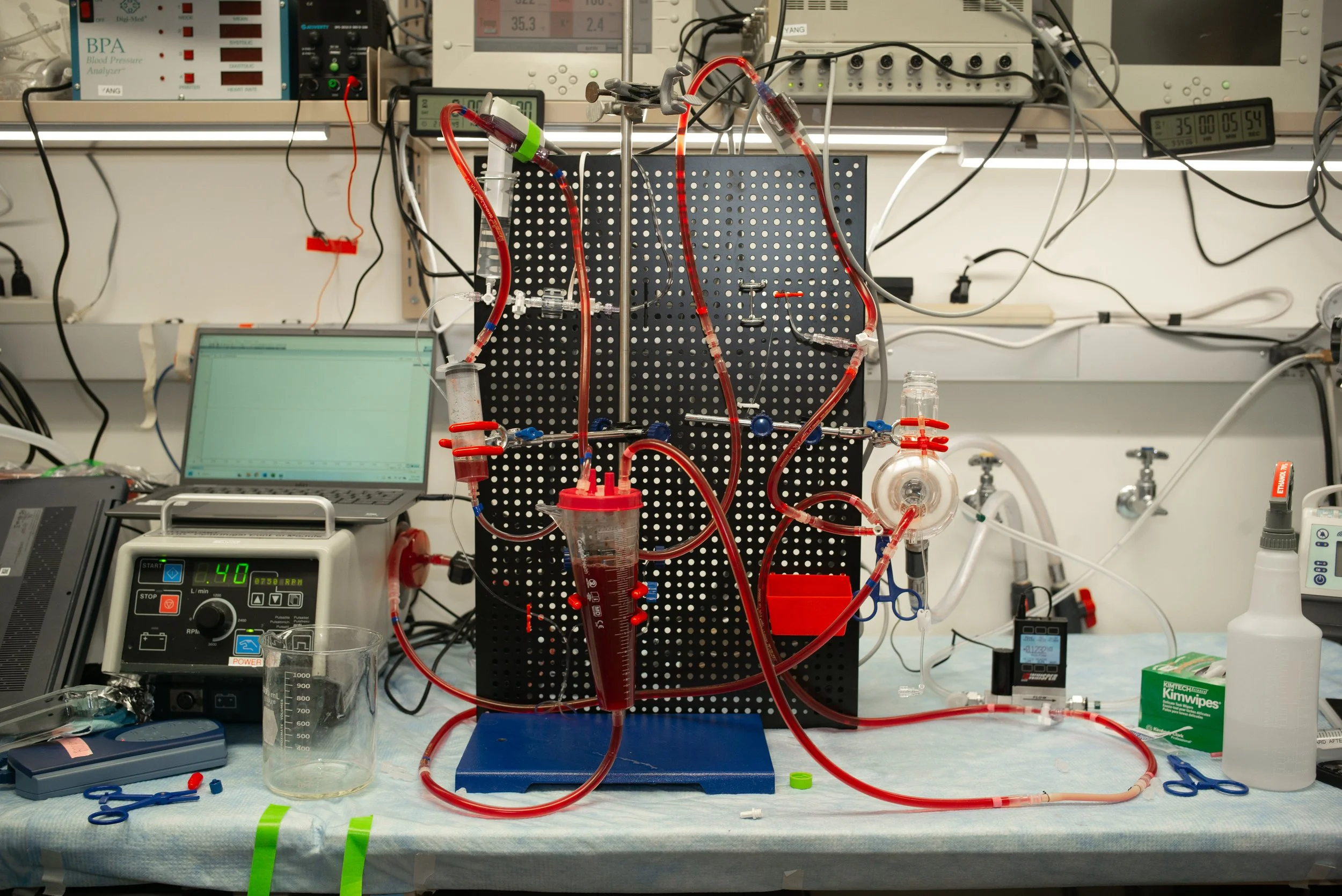 A laboratory setup with various scientific instruments, including a black pegboard with glass tubes, tubing, and clamps connected in a complex arrangement, surrounded by electronics, a laptop, measuring devices, and tools on a workbench.