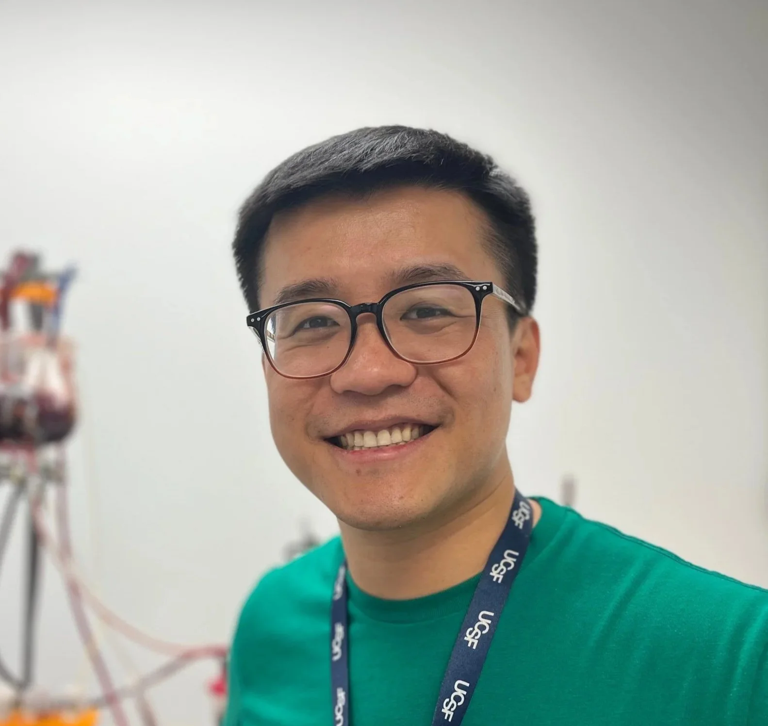 Young man wearing glasses and a lanyard with 'USCF' logo, smiling in a bright indoor space with scientific equipment in the background.