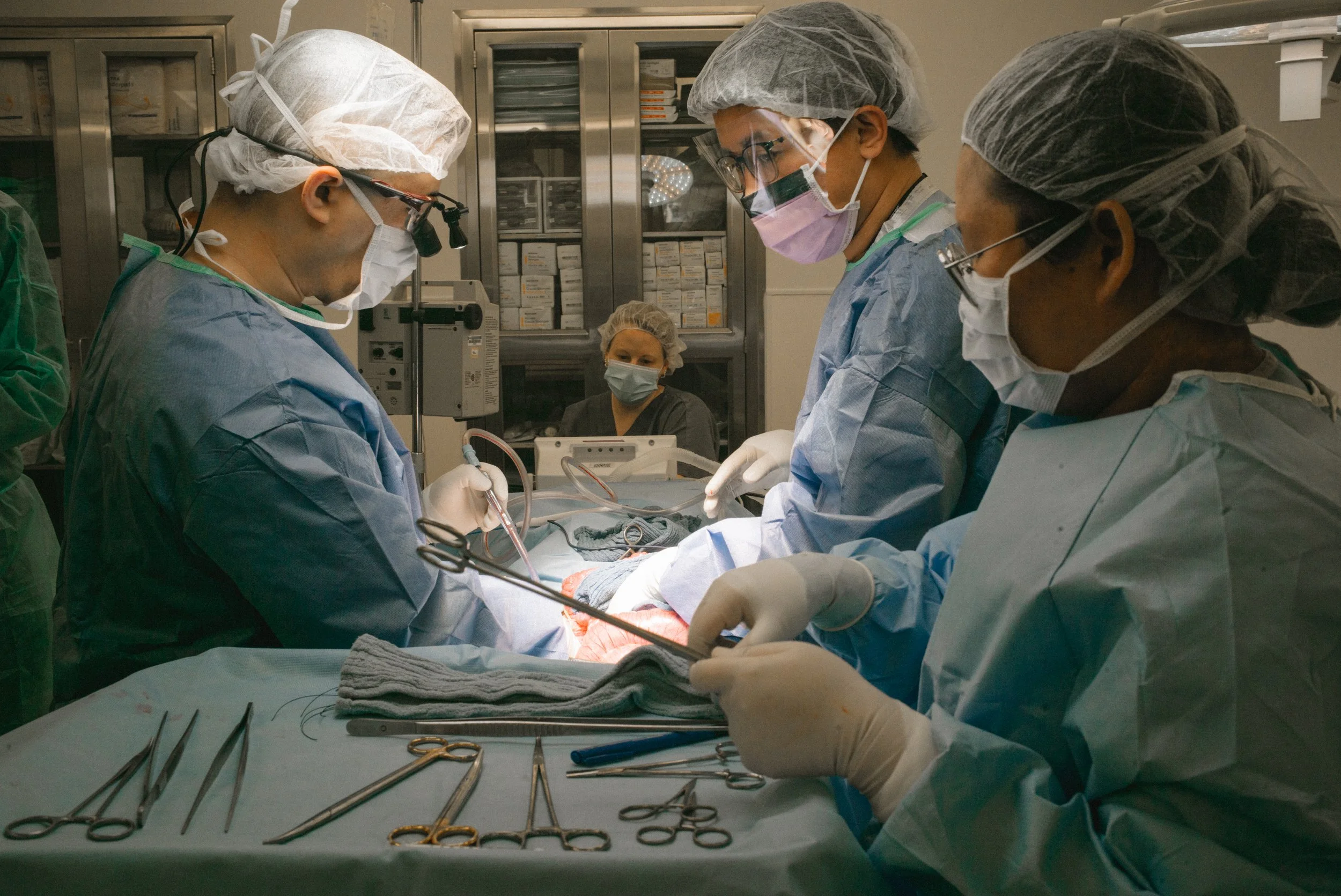 A team of surgeons performing an operation in an operating room, wearing surgical masks, gloves, and scrubs, with surgical instruments laid out on a table in the foreground.