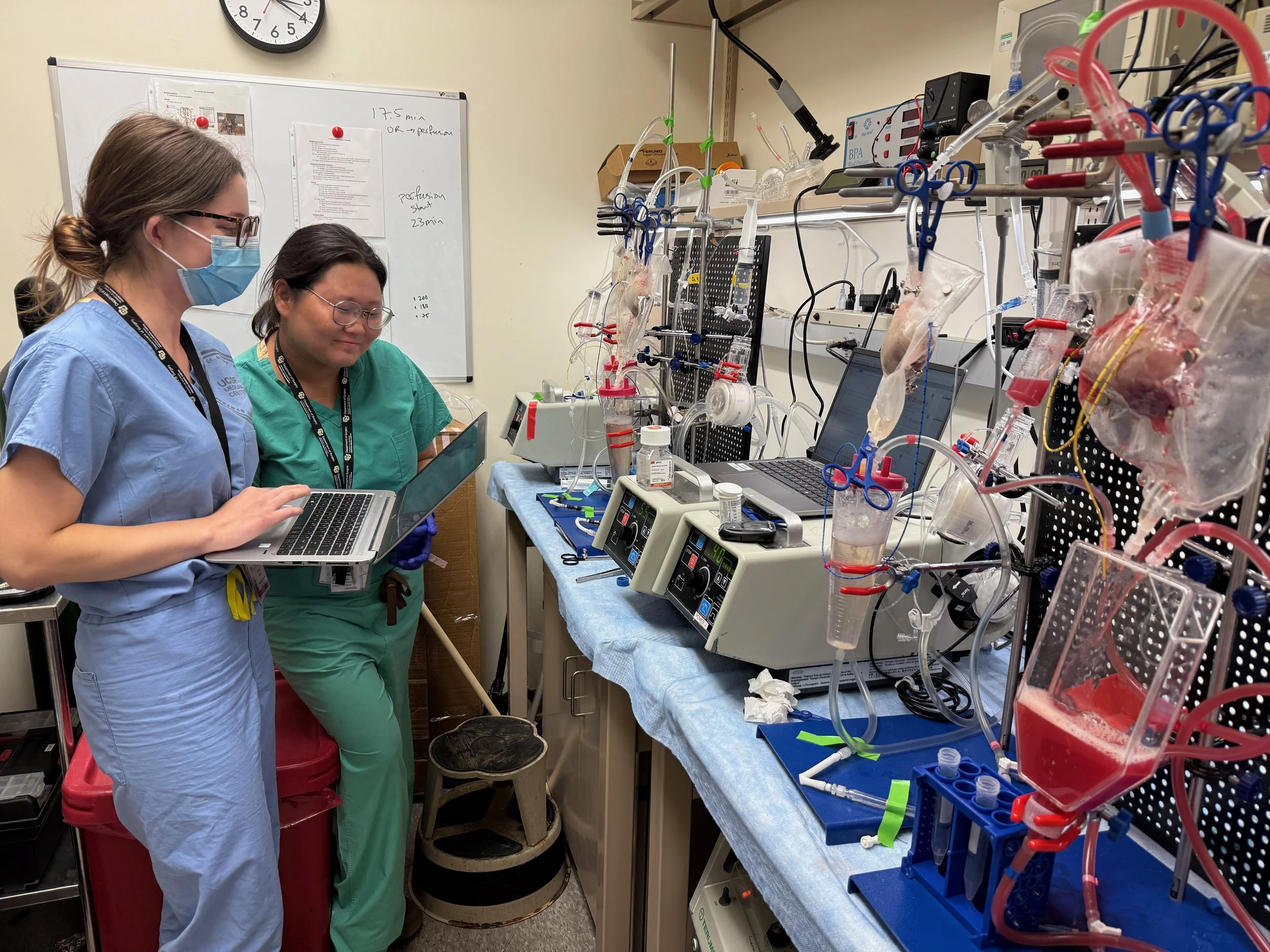 Two medical professionals, one wearing blue scrubs and a face mask, the other in green scrubs, working in a laboratory with advanced medical equipment, tubes, and computers.