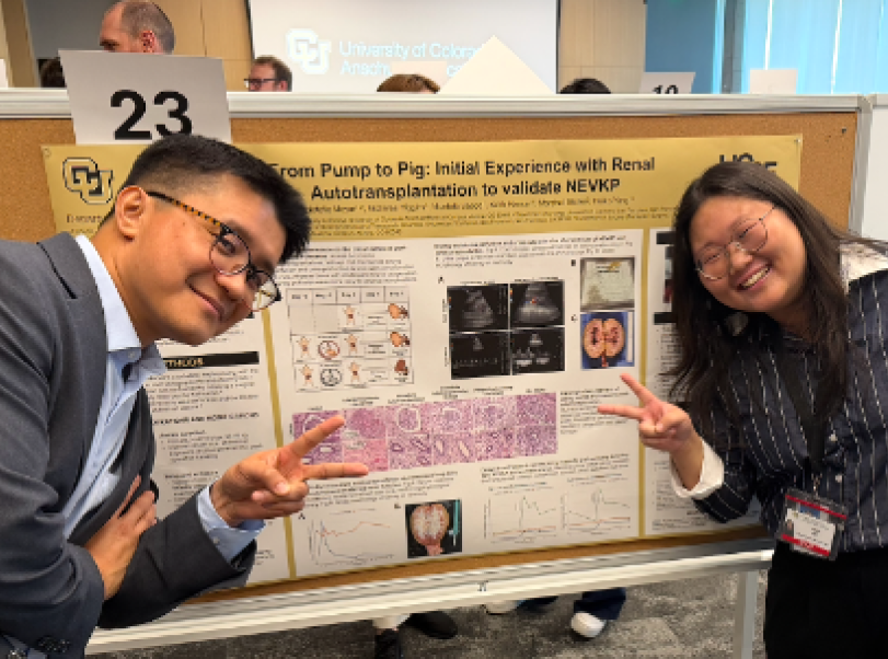 Two smiling individuals, a man and a woman, pose in front of a scientific research poster at a conference. Both are making peace signs with their hands. The poster contains text, images, and graphs, and the setting appears to be an academic or conference environment.