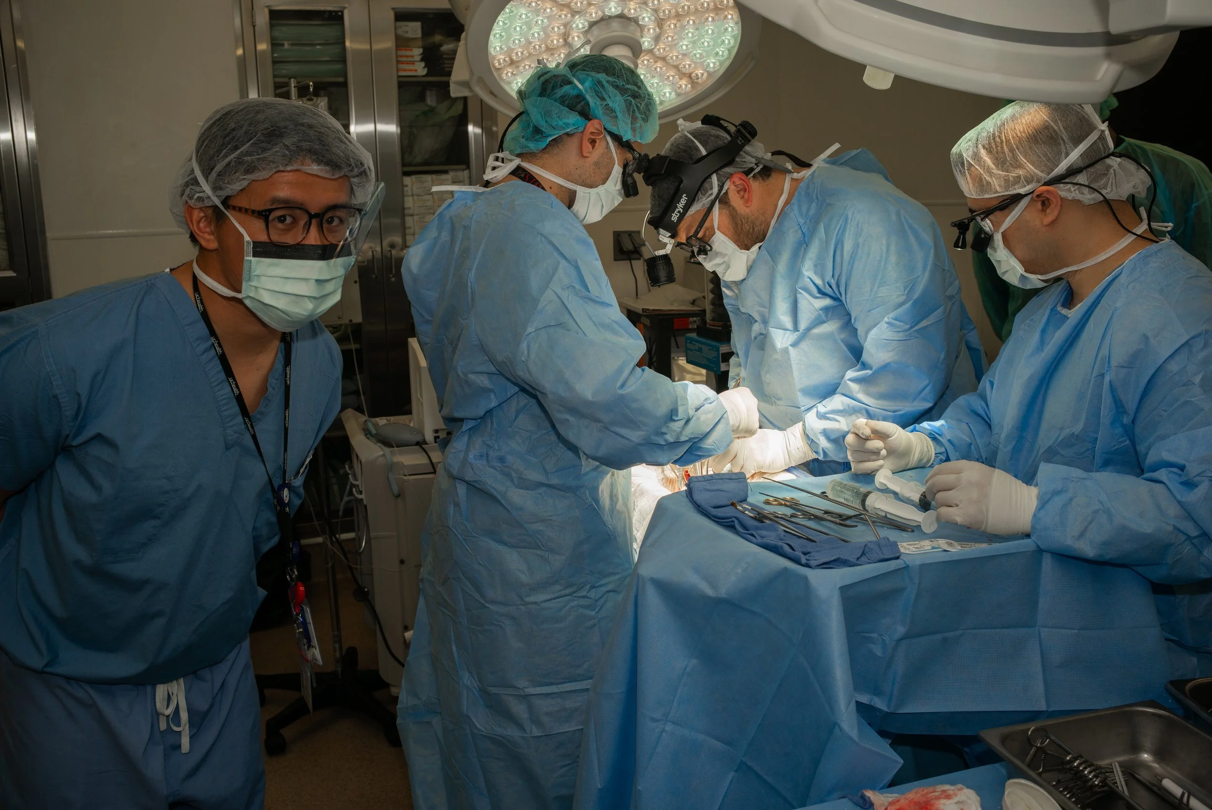 Medical team performing surgery in an operating room with surgeons wearing blue surgical gowns, masks, gloves, and surgical loupes, and operating under bright surgical lights.