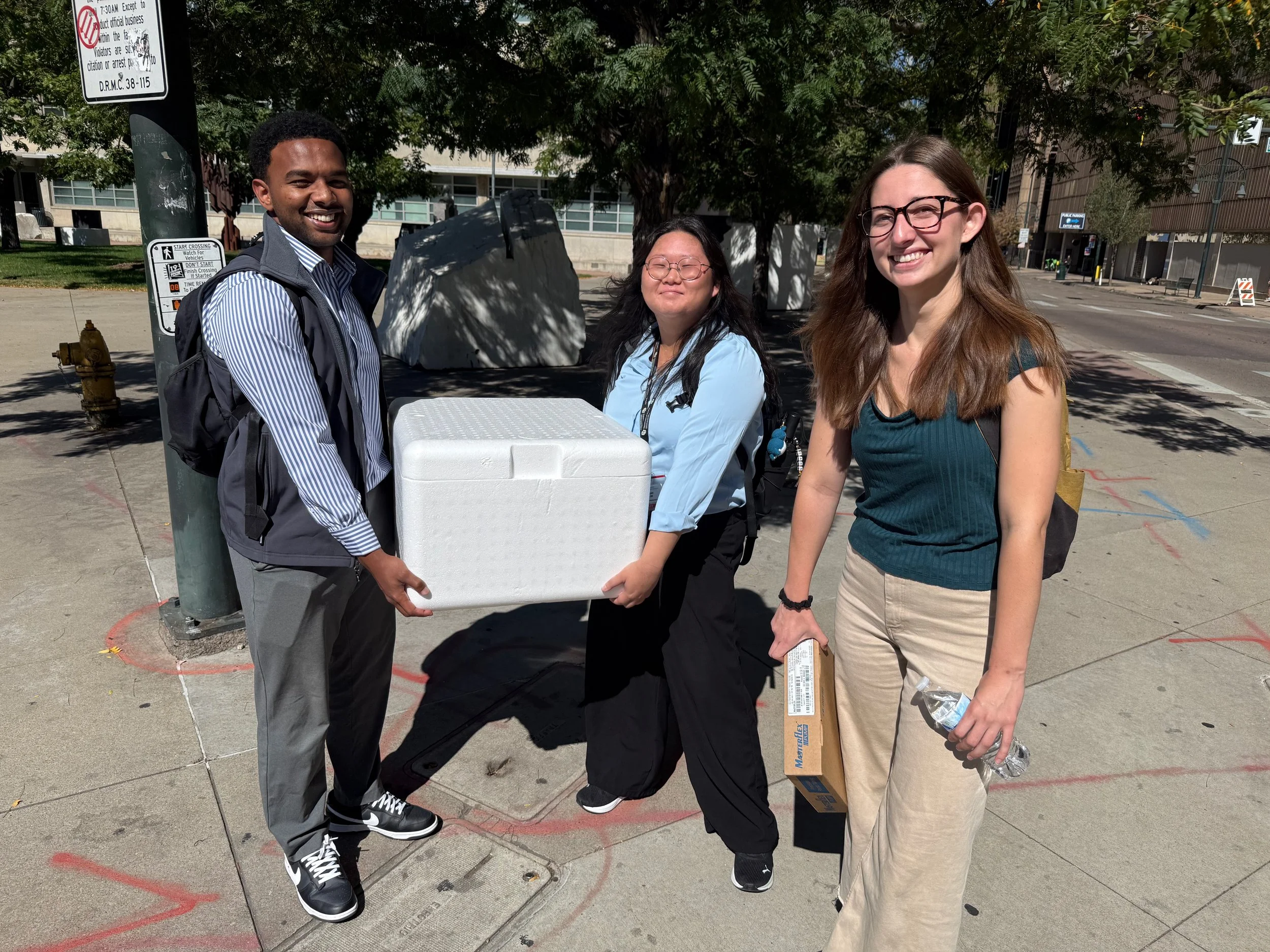 Three young adults standing outdoors on a sidewalk, holding a white styrofoam food container and a cardboard box, smiling at the camera. Two women and one man, with a tree and city buildings in the background.