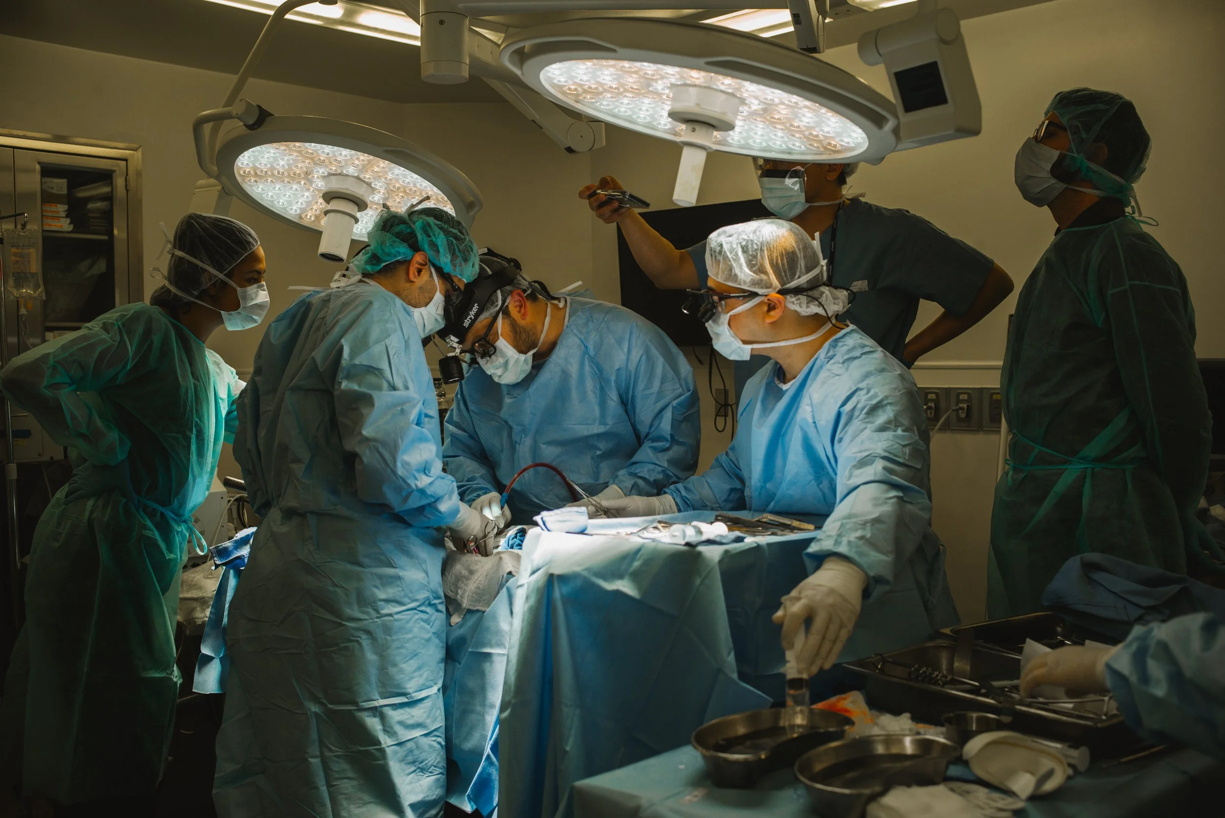 Medical team performing surgery in an operating room, wearing surgical gowns, masks, and gloves, with overhead surgical lights illuminating the procedure.