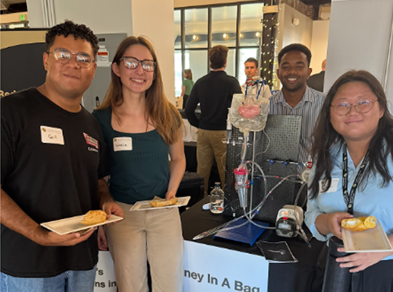 Four people standing together at a science fair or exhibition, smiling. They are holding plates with baked goods. Behind them is a display with scientific equipment, likely a scientific experiment or project, and other people in the background.