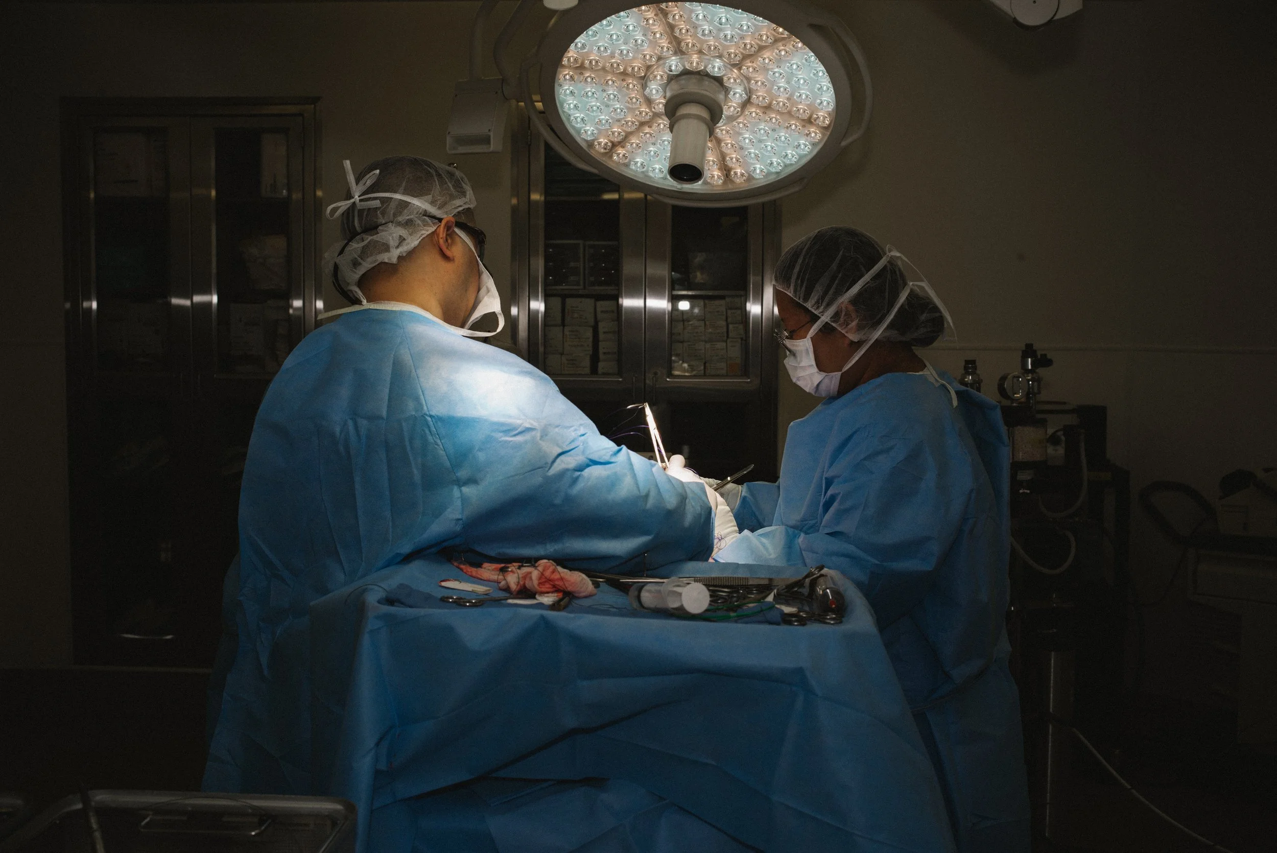 Two surgeons in blue gowns, masks, and caps performing surgery under a bright surgical light in an operating room.