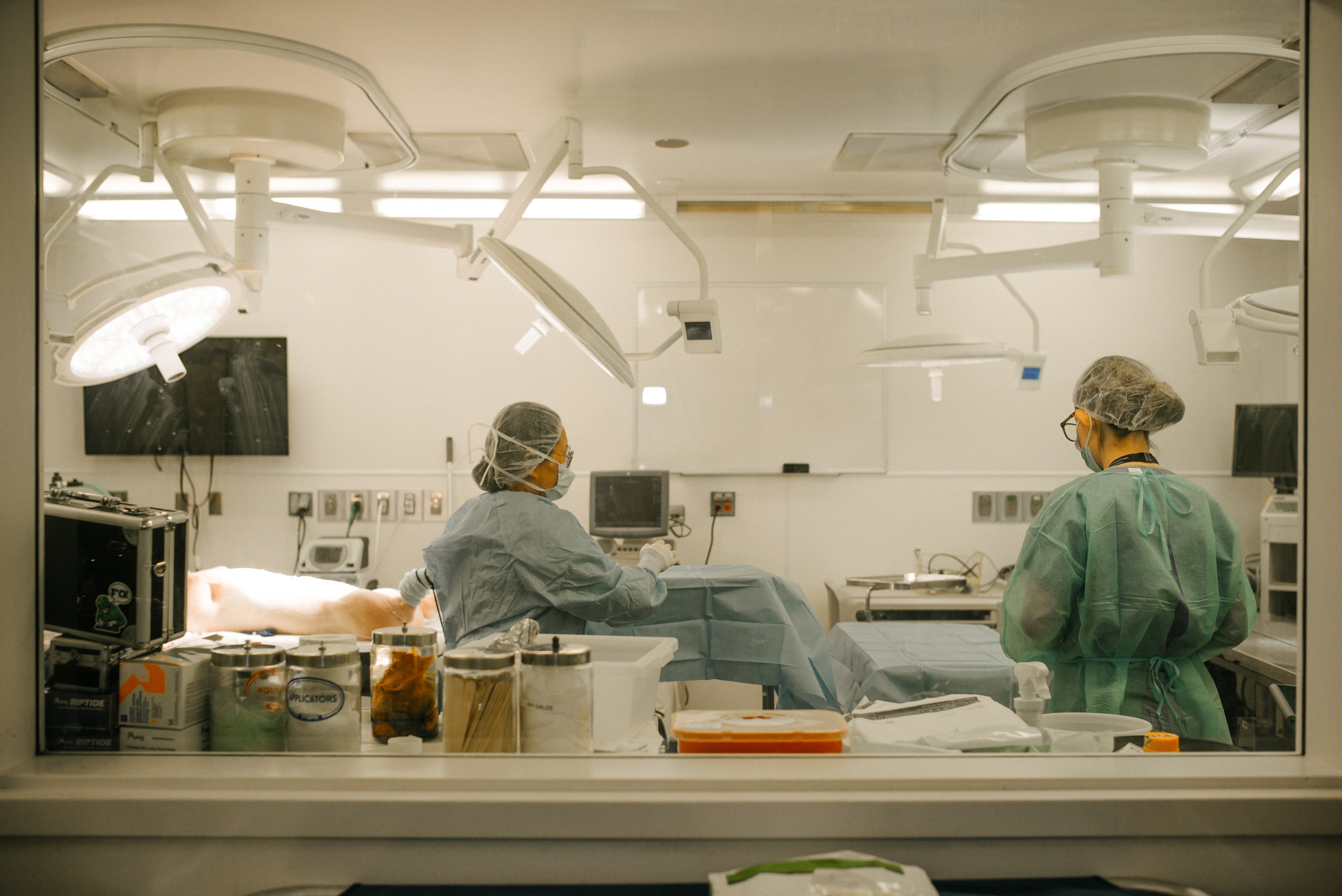 Medical professionals in surgical gowns, masks, and hair covers attending to a patient in an operating room with surgical lights and medical equipment.