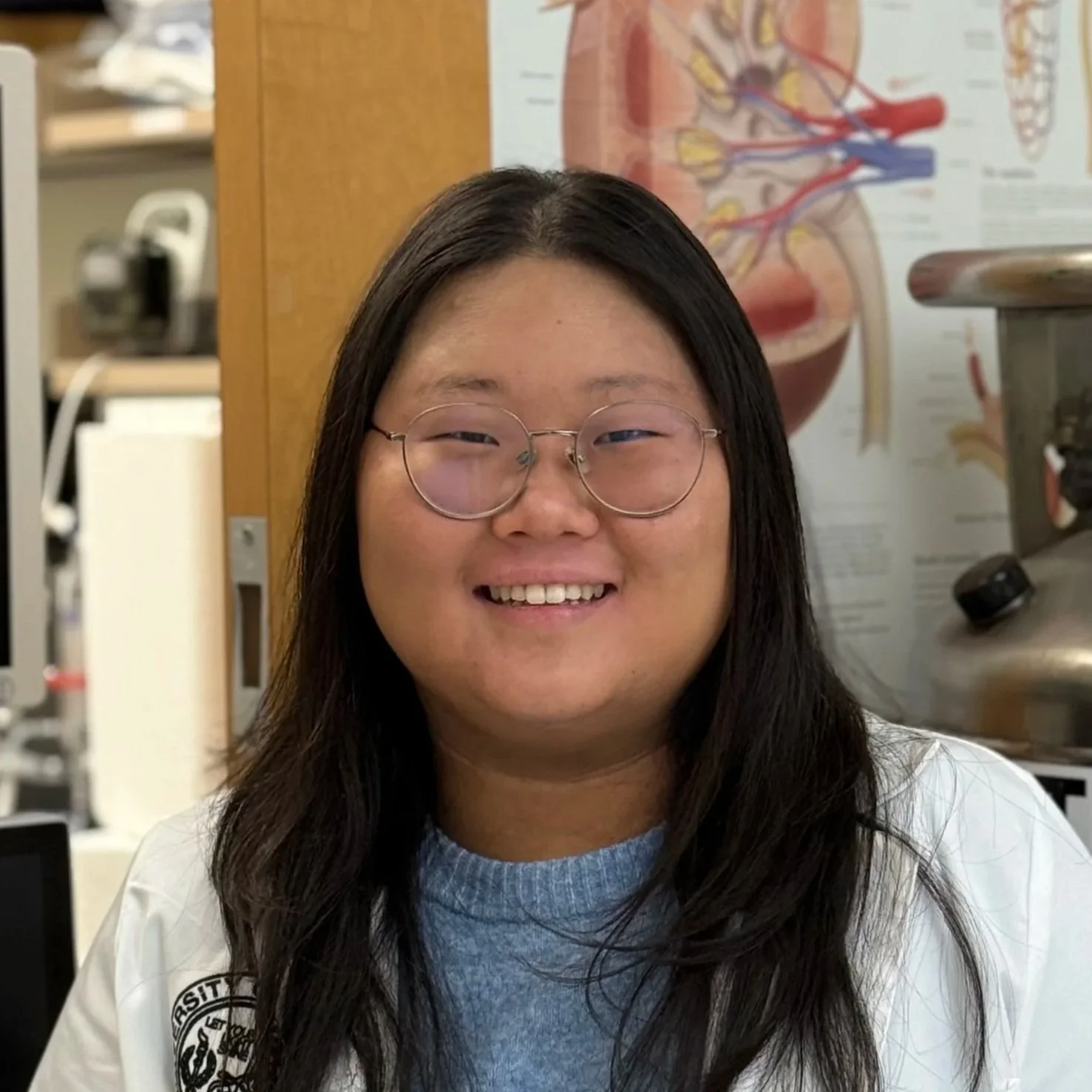 A smiling young woman with long dark hair and glasses in a medical or laboratory setting, with anatomical posters and equipment in the background.