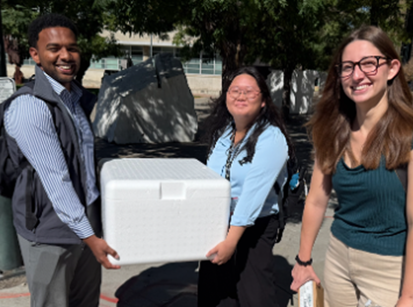 Three people smiling outdoors, one person holding a white container shaped like a cooler, with trees and a building in the background.