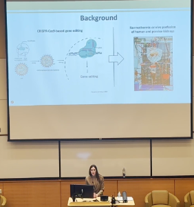 A woman giving a presentation in a classroom or lecture hall, with a large slideshow projected on the screen behind her showing diagrams and text on a background about coronavirus and immune response.