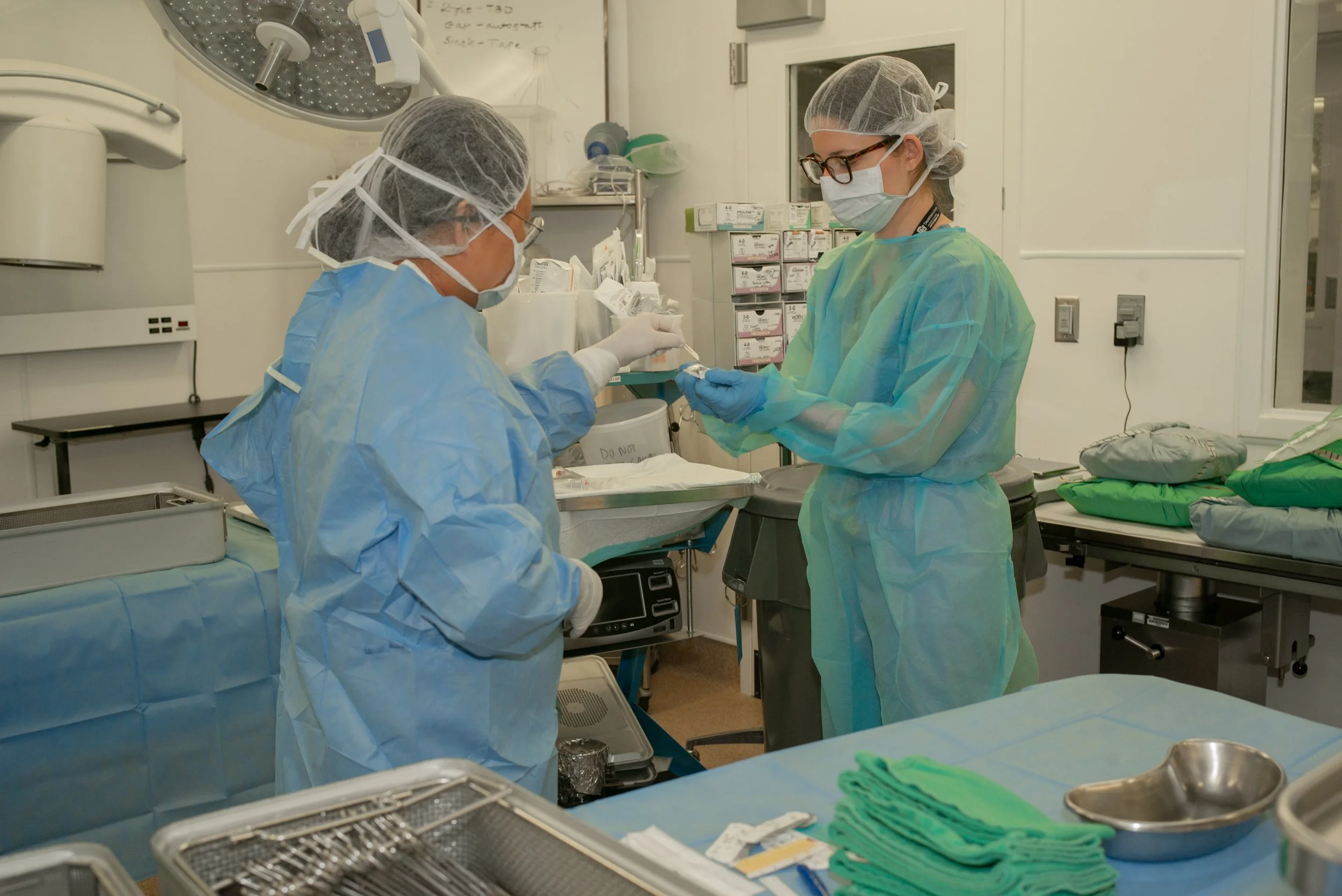 Two healthcare professionals in scrubs, masks, gloves, and hairnets preparing a medical syringe in an operating room or clinical setting.