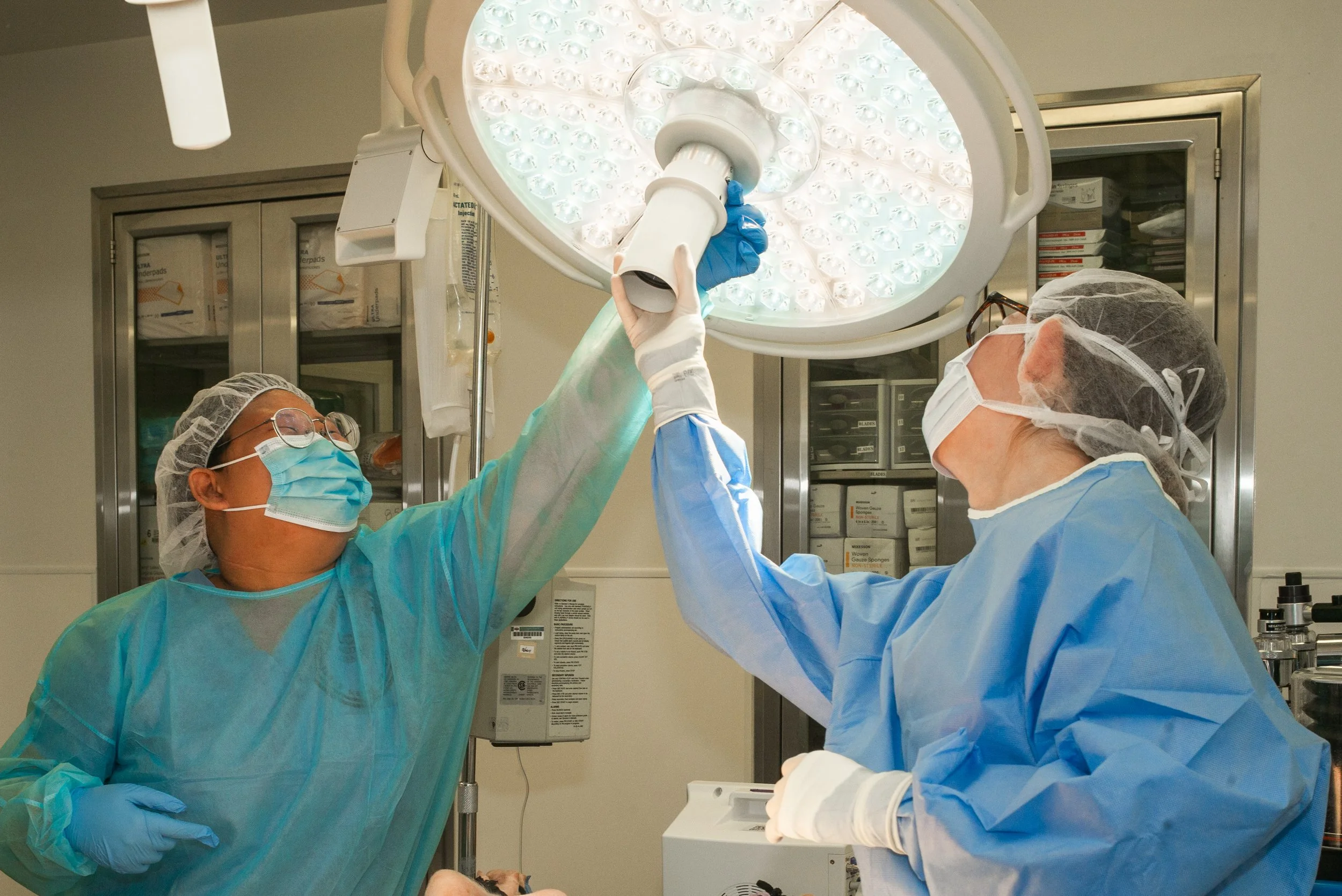 Two medical professionals in protective gear, masks, and gloves working under an overhead surgical light in an operating room.