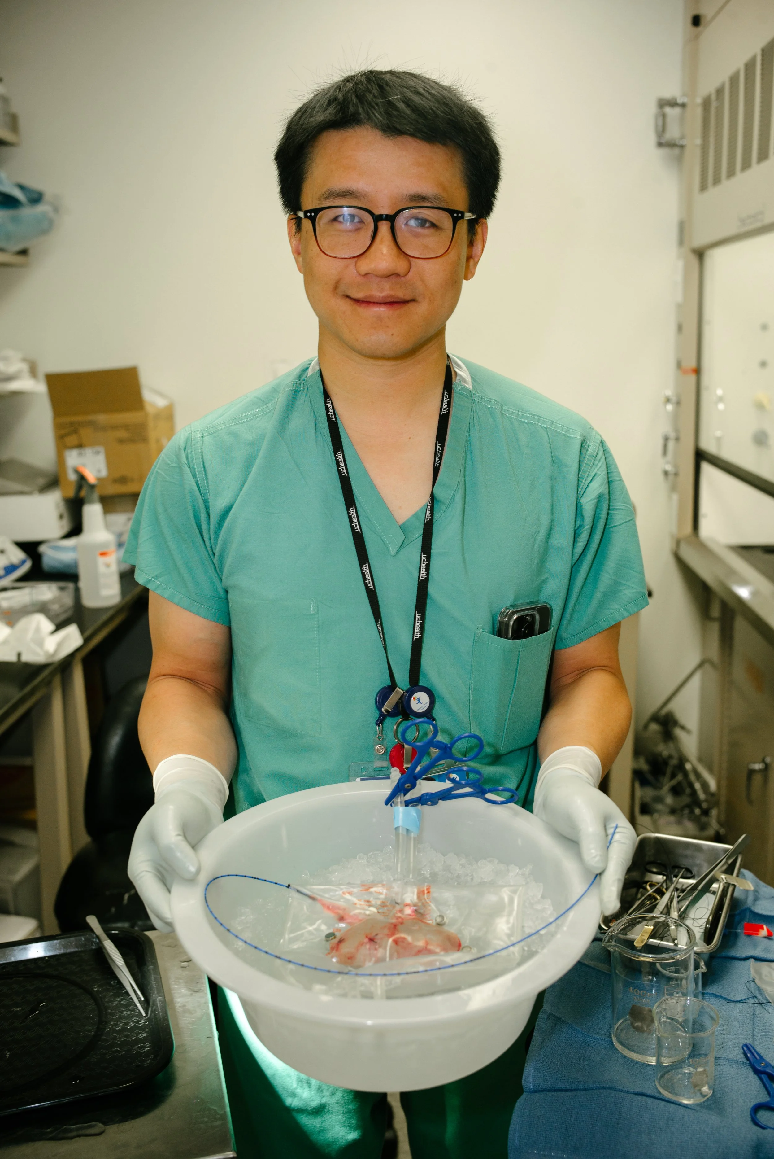 A medical professional in green scrubs and glasses holding a white basin with a small animal inside, surrounded by medical tools, in a laboratory or clinical setting.