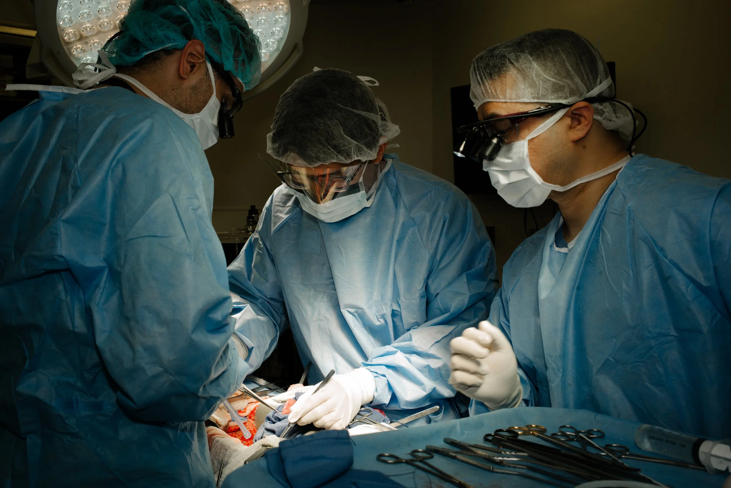 Medical professionals in surgical gowns, masks, and gloves working together in an operating room under bright surgical lights.