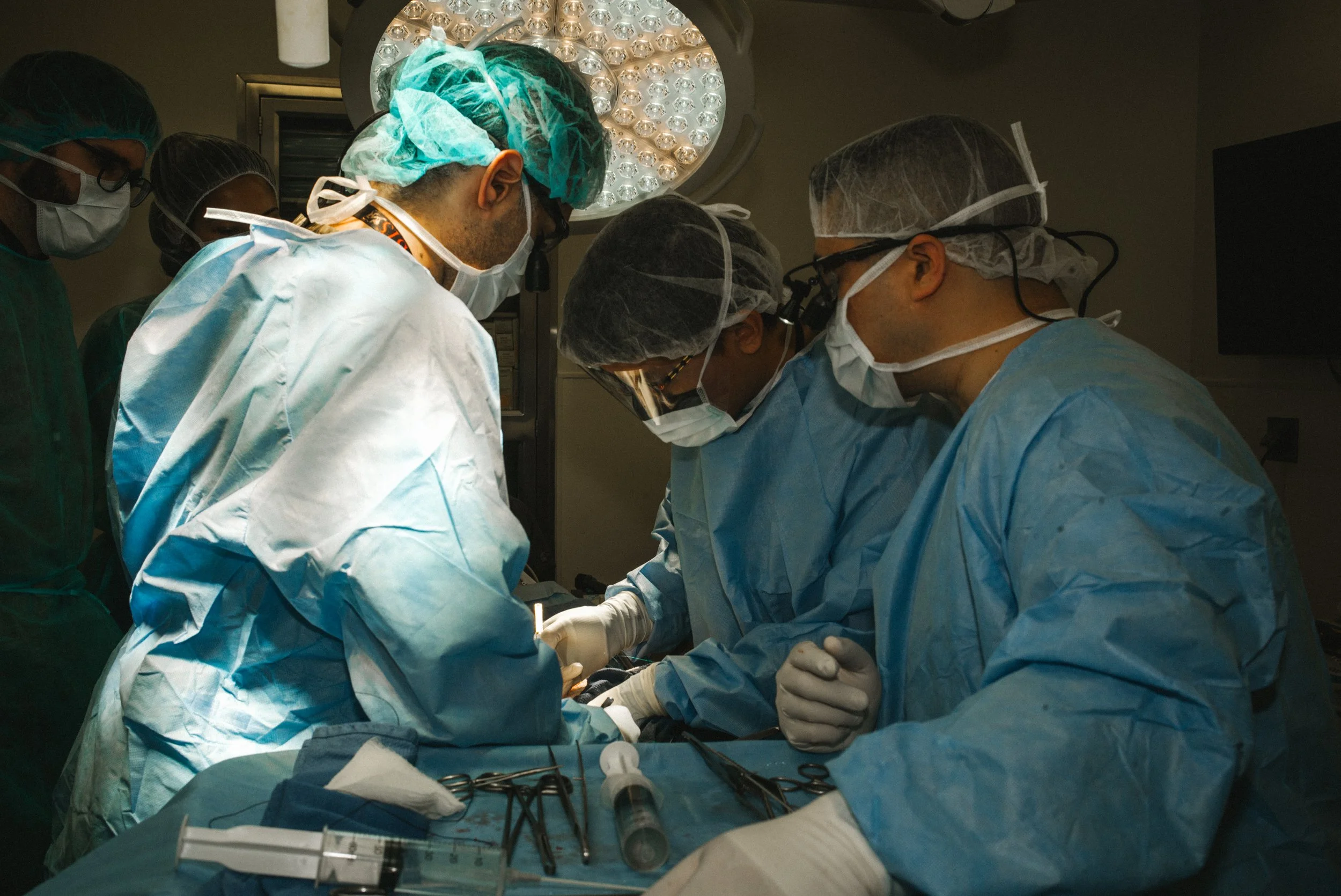 A group of surgeons in scrubs and masks performing surgery in an operating room, working closely around the patient.