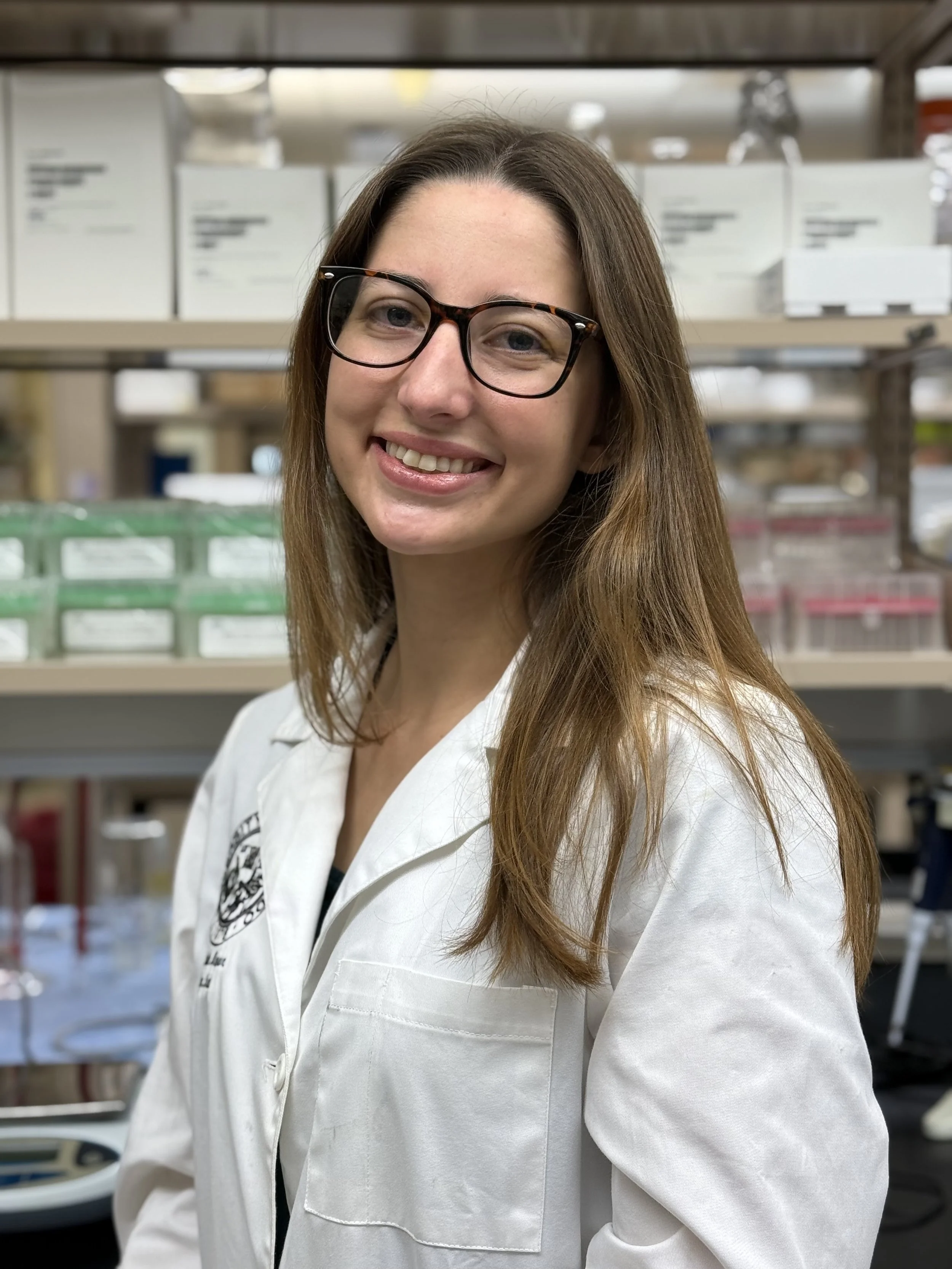 A woman with glasses and long brown hair smiling at the camera in a laboratory setting.