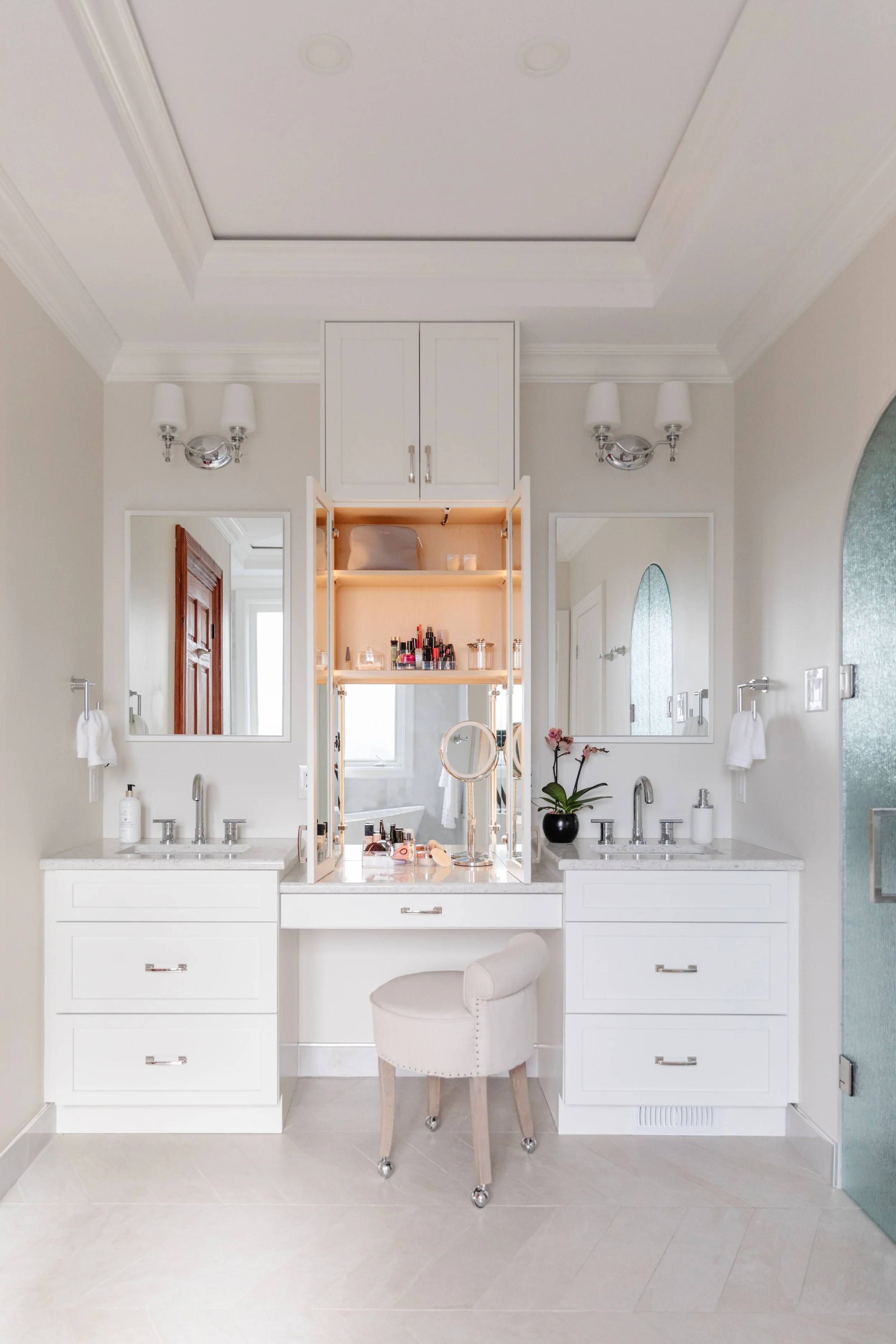 A bright, elegant bathroom with a central vanity area featuring a large mirror, two sinks, and a padded stool. The vanity has open storage with shelves holding beauty products. Two wall-mounted mirrors and sconces provide lighting. The room has white cabinetry, a light-colored tiled floor, and decorative crown molding with a coffered ceiling.