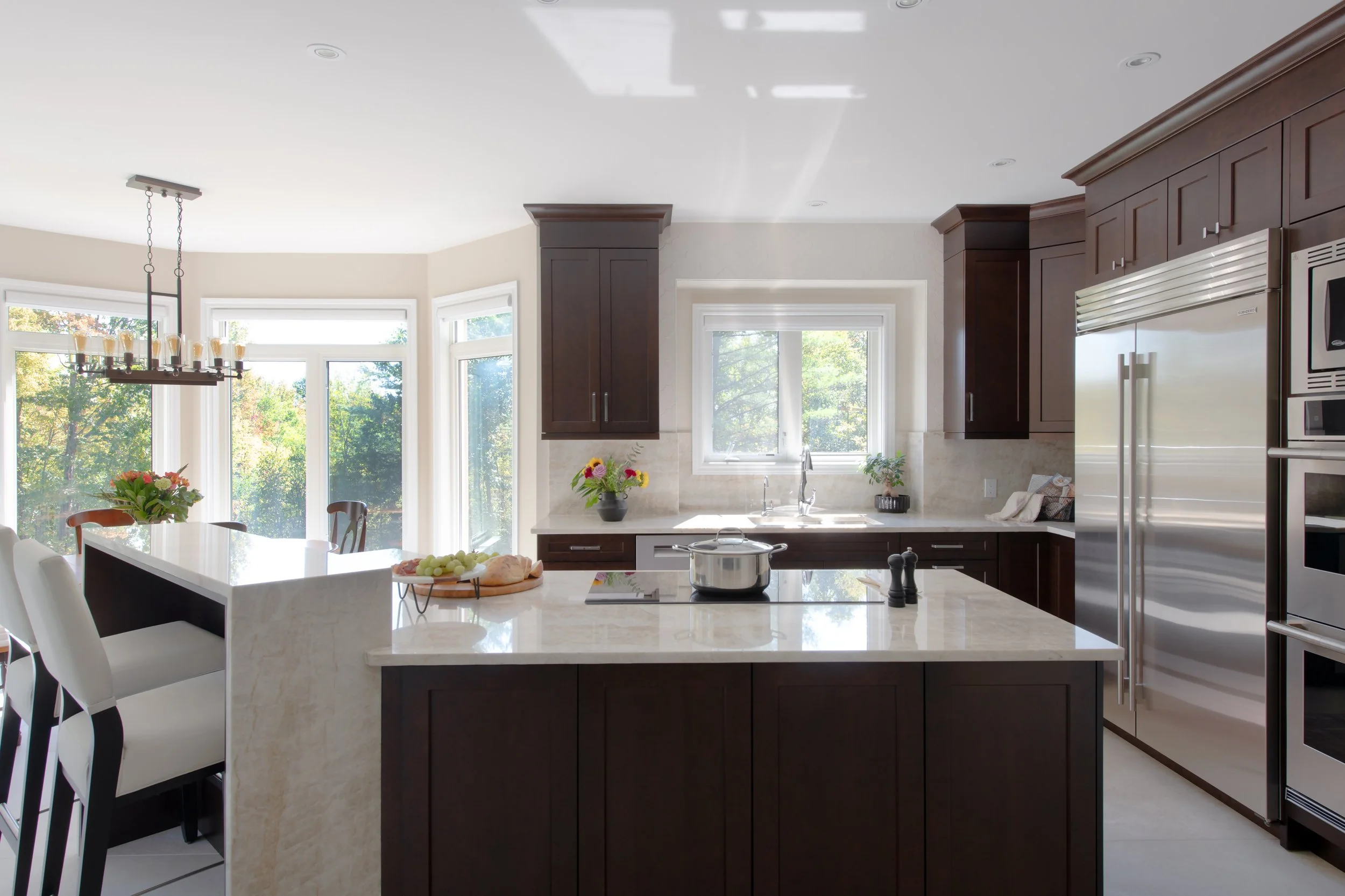 Bright kitchen with dark wood cabinets, white countertops, stainless steel appliances, and large windows showing a garden view.