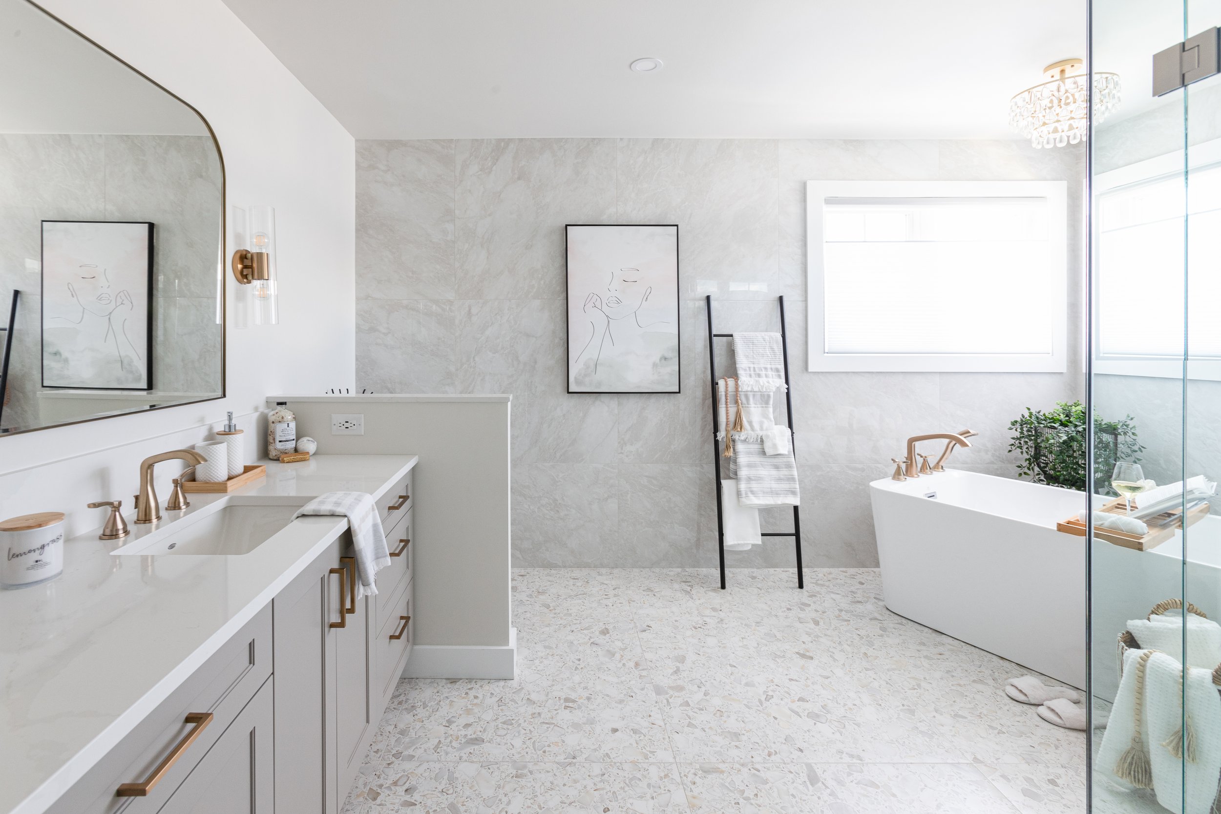 Modern bathroom with light-colored walls and flooring, featuring a large mirror above a white vanity with gold hardware, a bathtub with a glass partition, and a ladder towel rack near a window, decorated with minimalistic art and decor.