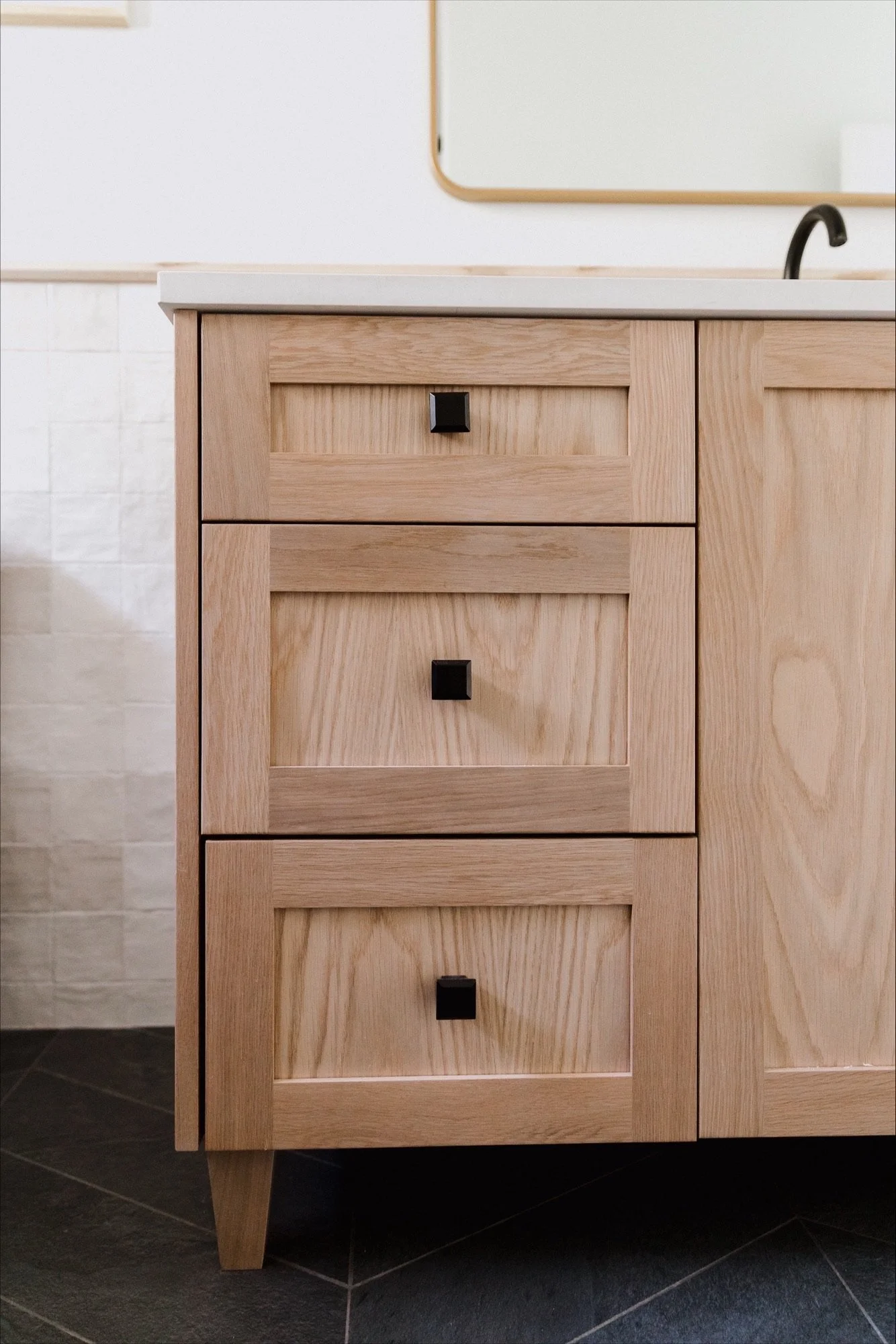 A minimalist bathroom with white walls, a white toilet with a decorative vase with black branches on top, a white textured wastebasket, and part of a vanity with drawers, a mirror, and a wall-mounted light fixture.