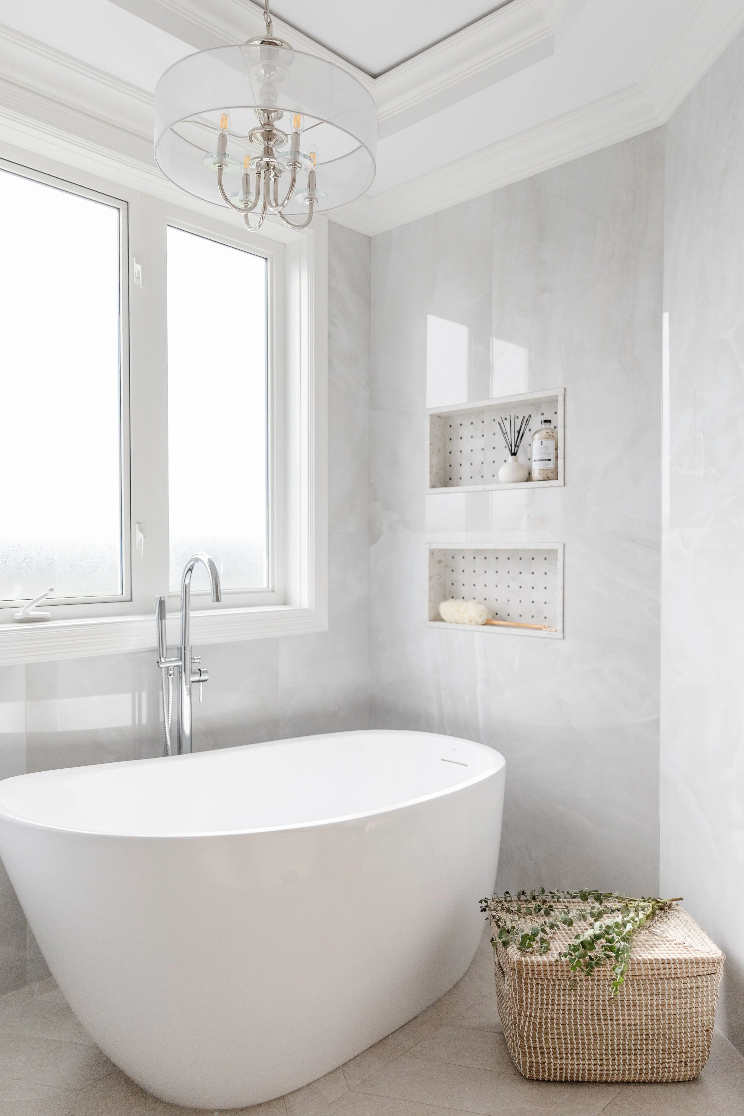 A modern bathroom with a white freestanding bathtub beneath a frosted window, a silver chandelier hanging from the ceiling, and built-in wall shelves for decor. A wicker basket with greenery is on the floor.