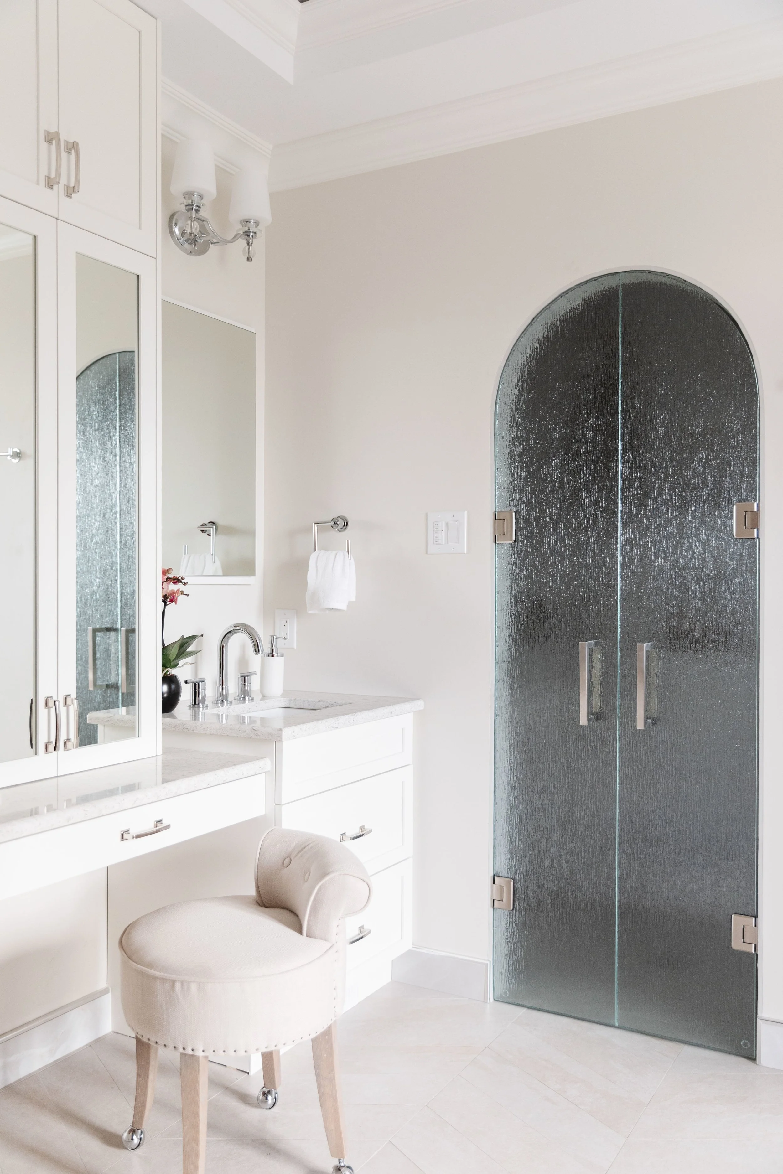 A bathroom with white cabinetry, a small upholstered chair, a glass shower door, and minimal decor.