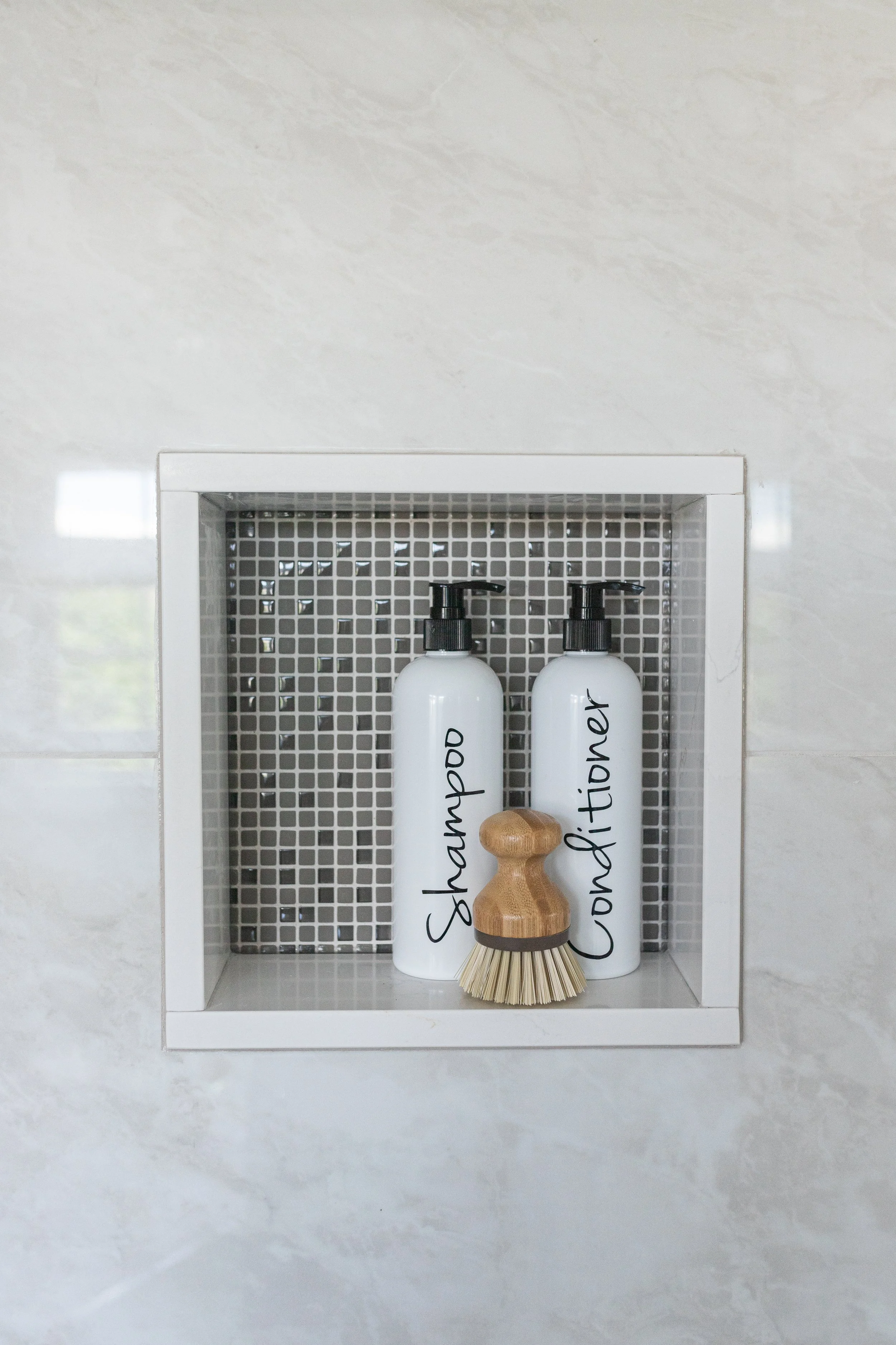 Shampoo and conditioner bottles labeled on a shower niche wall, with a scrubbing brush in front.