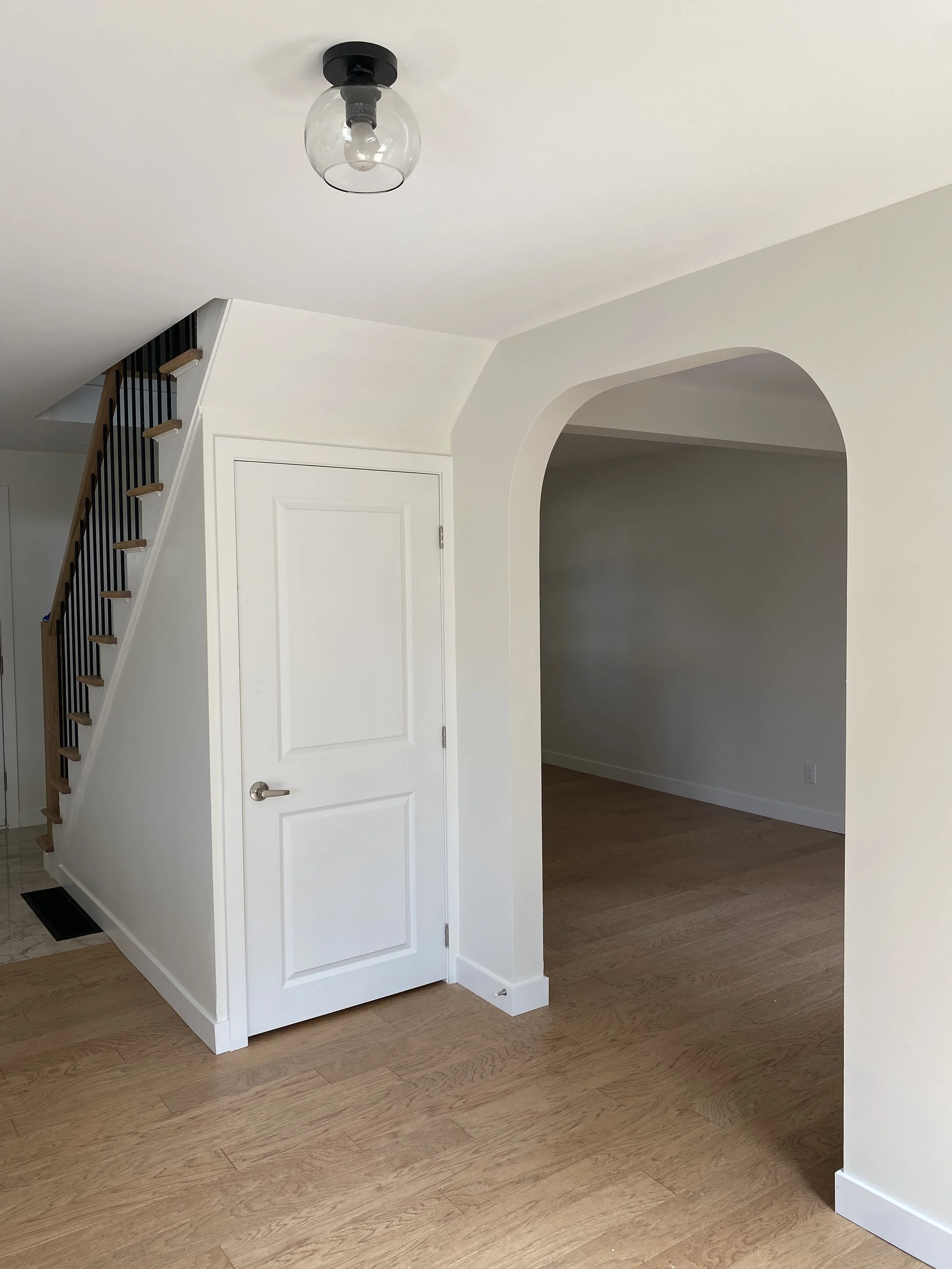 Bright bathroom with a white bathtub, glass-enclosed shower with a rain showerhead, and a window with white blinds. Decor includes a green plant, slippers, towels, a basket, and a wooden stool.