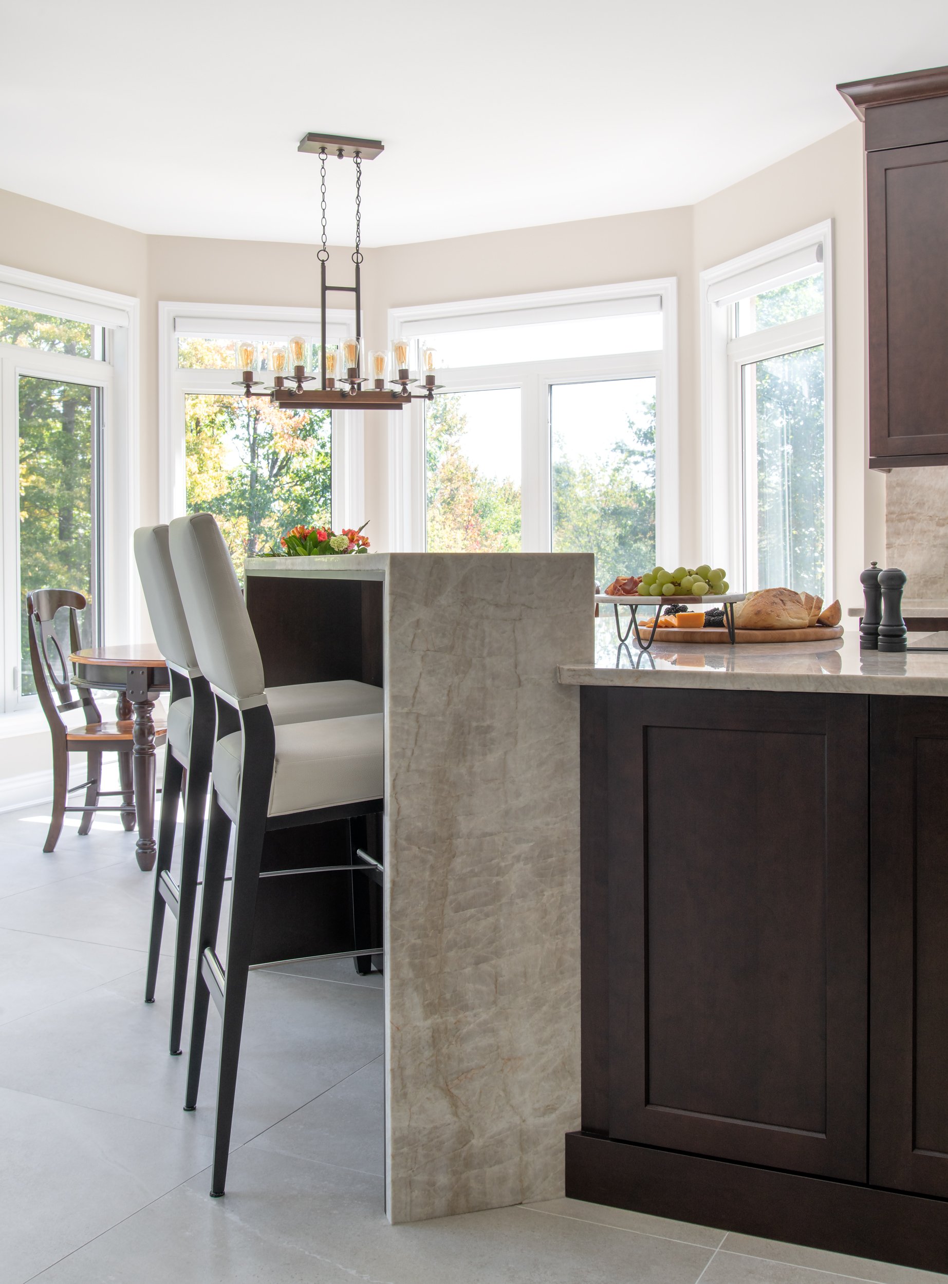 Bright kitchen with large windows, dark wood cabinets, marble counter with plates of grapes and bread, and high chairs.