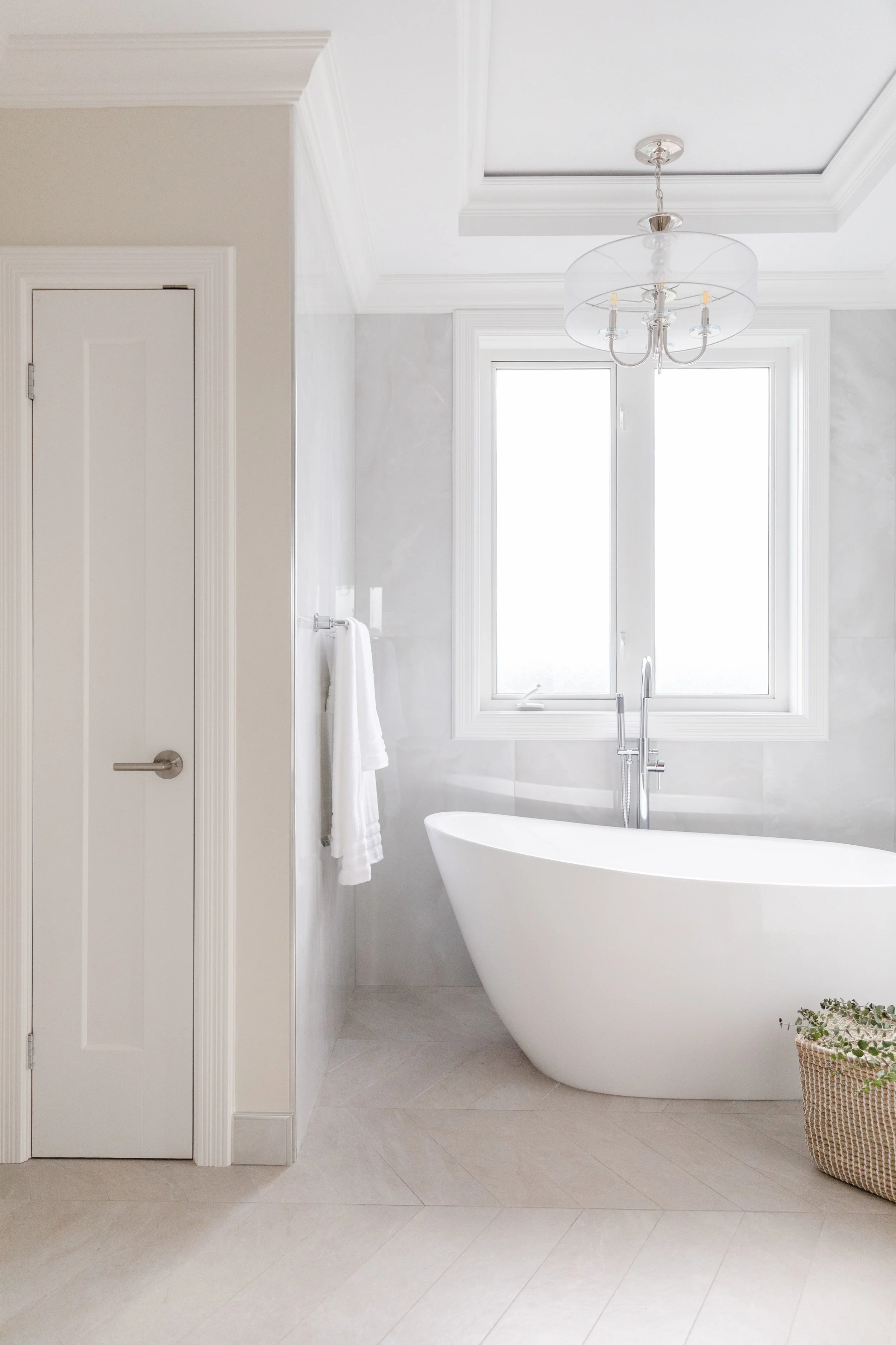 Modern bathroom with a freestanding white bathtub, large window, chandelier, towel, and basket with plants.