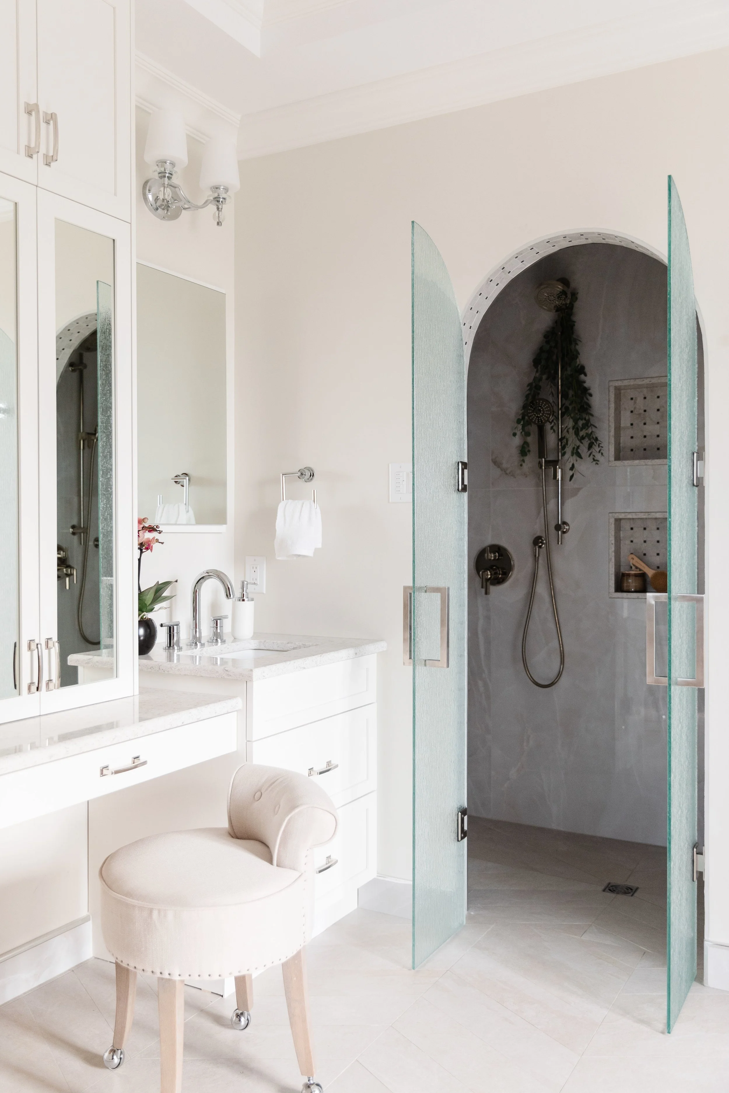 A bright, minimalist bathroom with a glass-enclosed shower, white vanity with a marble countertop, a large mirror, a beige upholstered stool, and a wall-mounted light fixture.