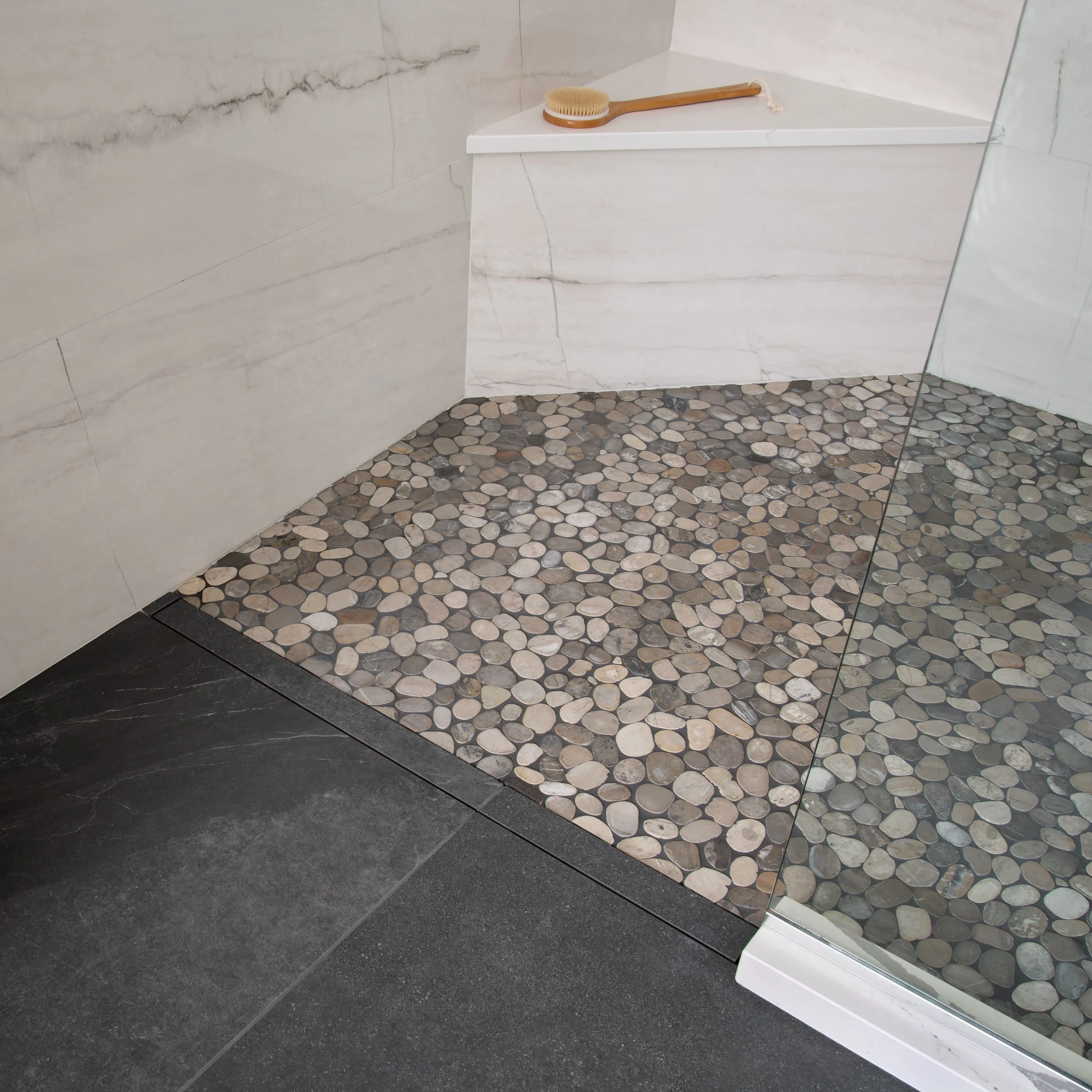 Corner of a modern shower with white marble walls, pebble stone shower floor, a glass partition, and a wooden scrub brush on a shelf.