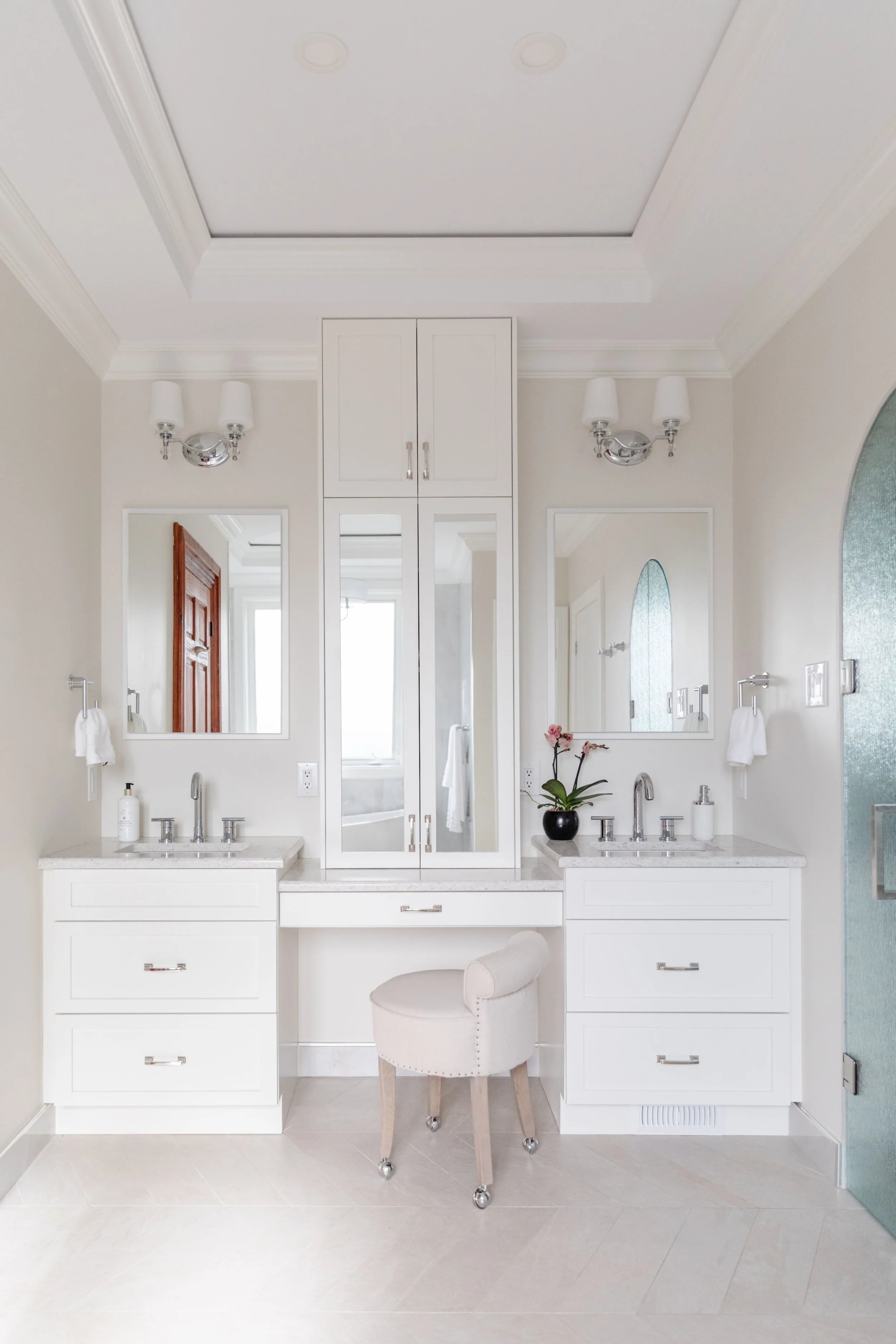 A bright, white bathroom vanity with two sinks, a central mirrored cabinet, and a stool. There are wall sconces, mirrors, and a black plant pot with pink orchids.