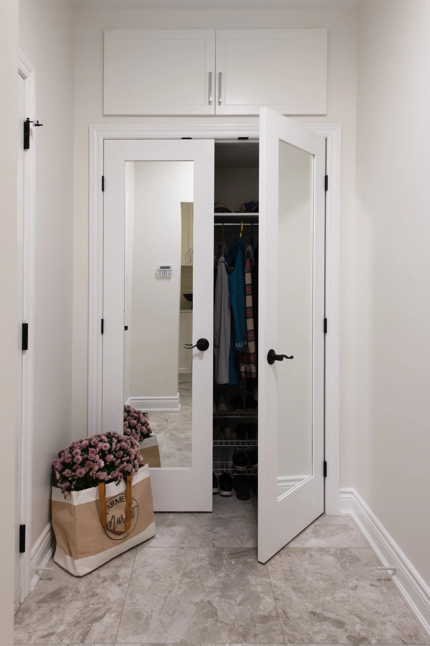 An open closet door revealing hanging clothes, a shoe rack, and a mirror reflecting a hallway. There is a bag with pink flowers and brown paper shopping bags on the floor beside the closet.