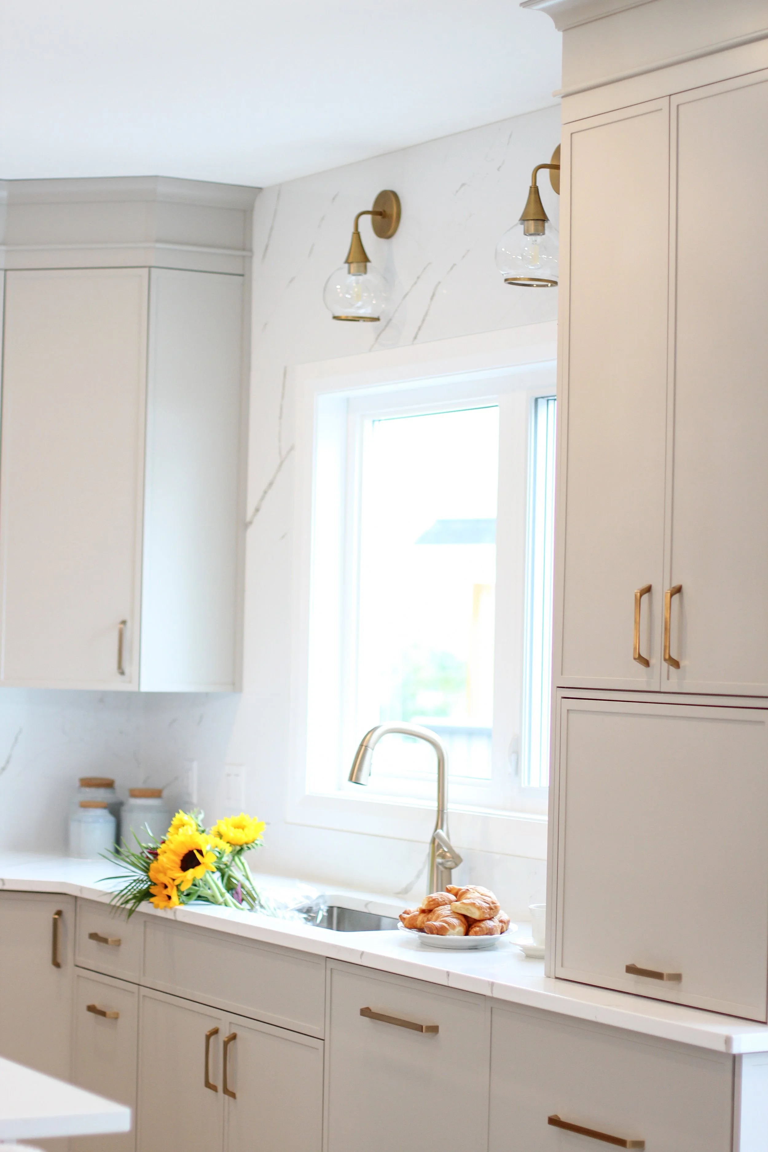 Bright kitchen with white cabinets, gold handles, a window, a sunflowers bouquet, and a plate of croissants on the countertop.