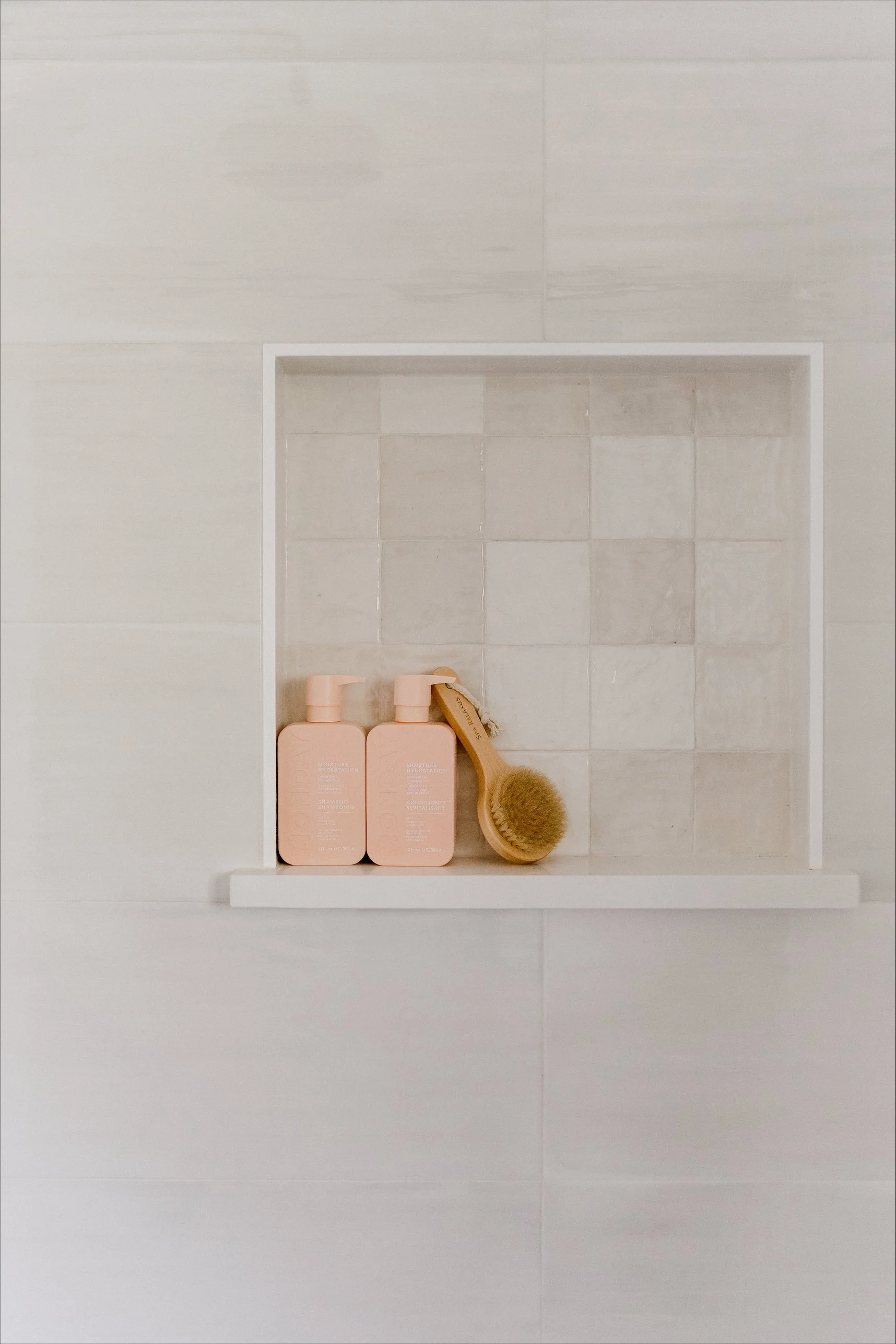 Shampoo and conditioner bottles labeled on a shower niche wall, with a scrubbing brush in front.