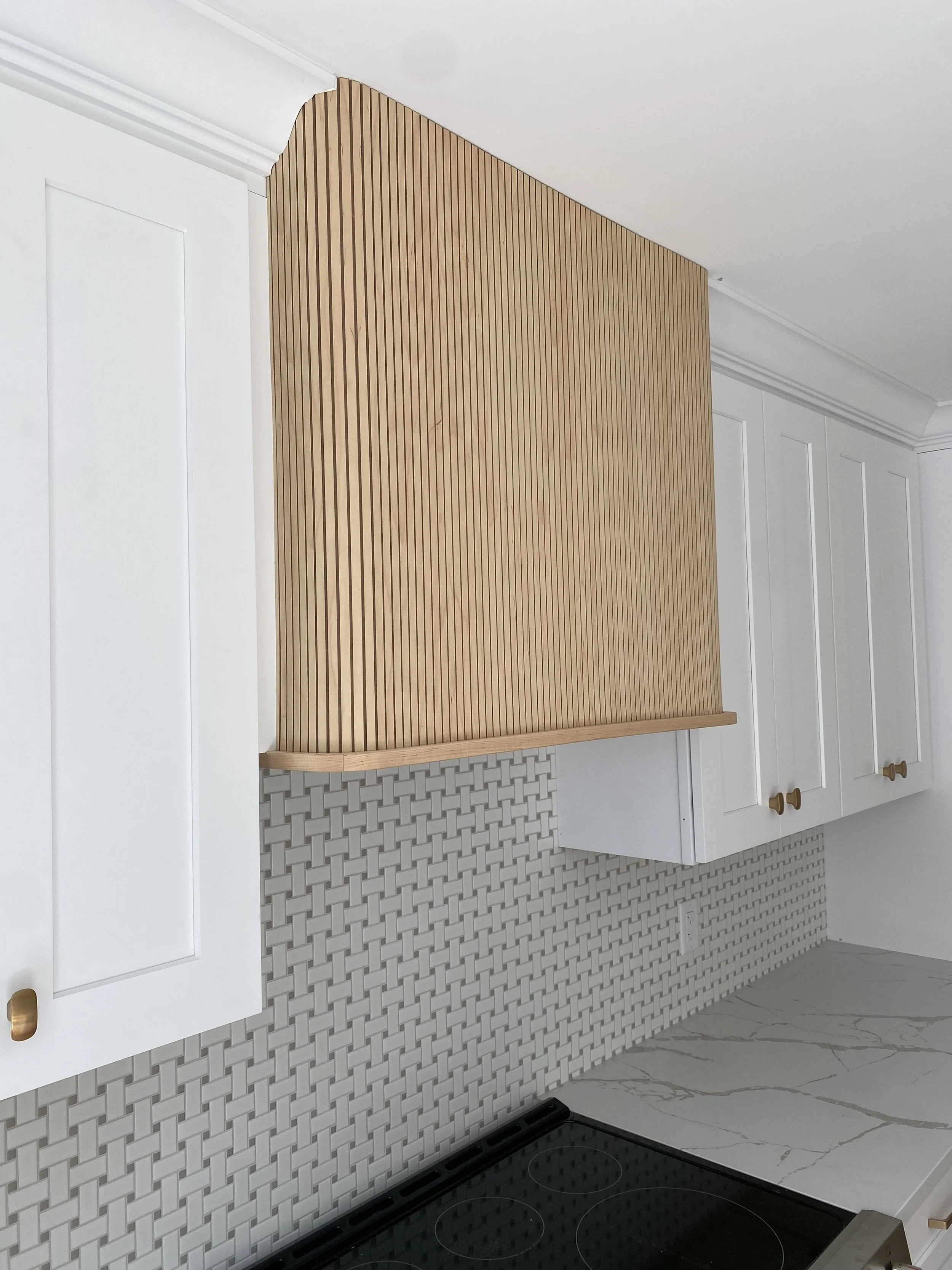 Shower with a brass shower head and fixtures, a built-in shelf with toiletries, a hanging plant, and a small wooden stool in a modern bathroom with light gray marble walls.