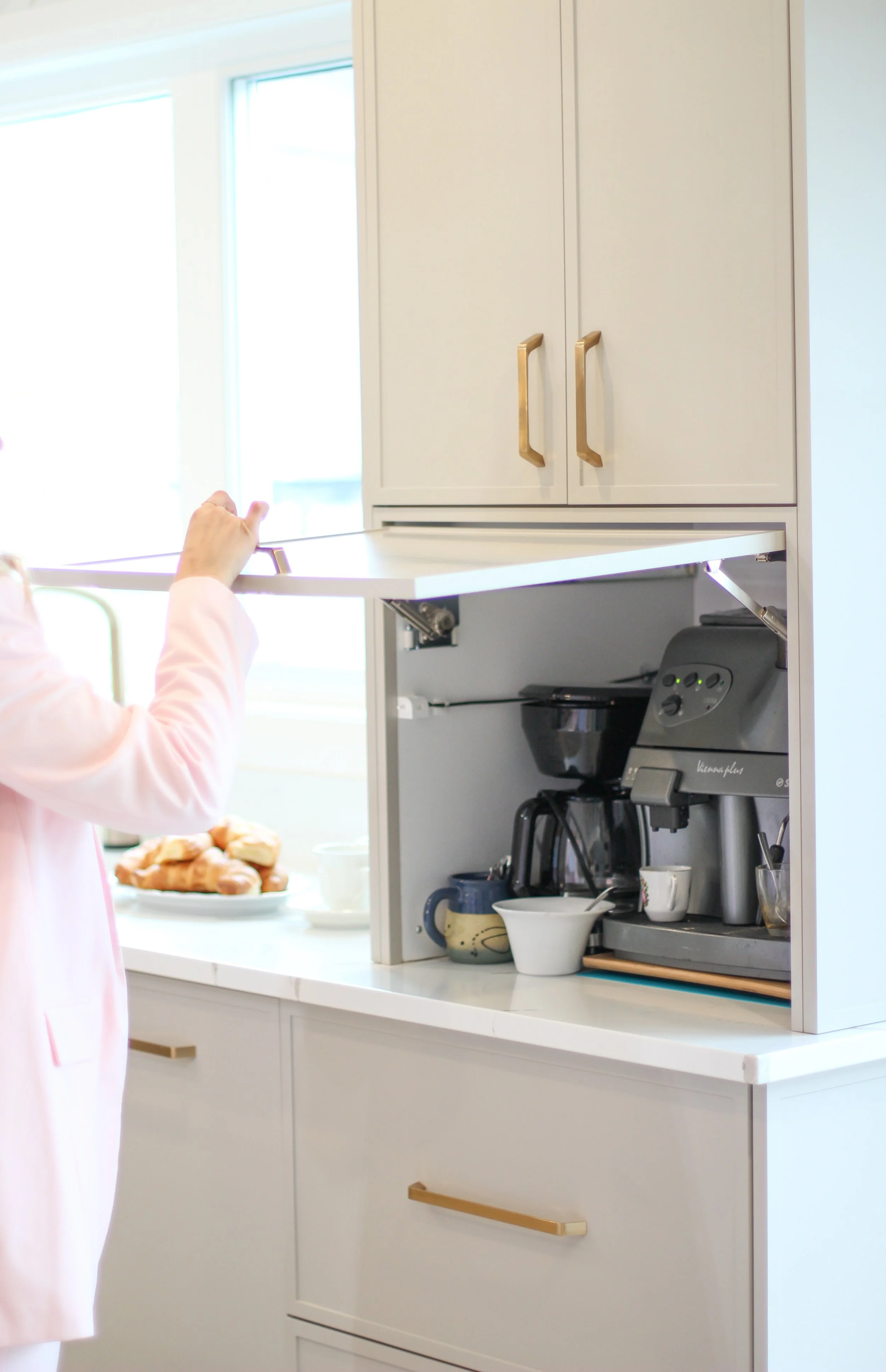 A person opening a cabinet door above a kitchen counter, revealing a coffee machine and various cups and mugs, with a plate of pastries in the background.