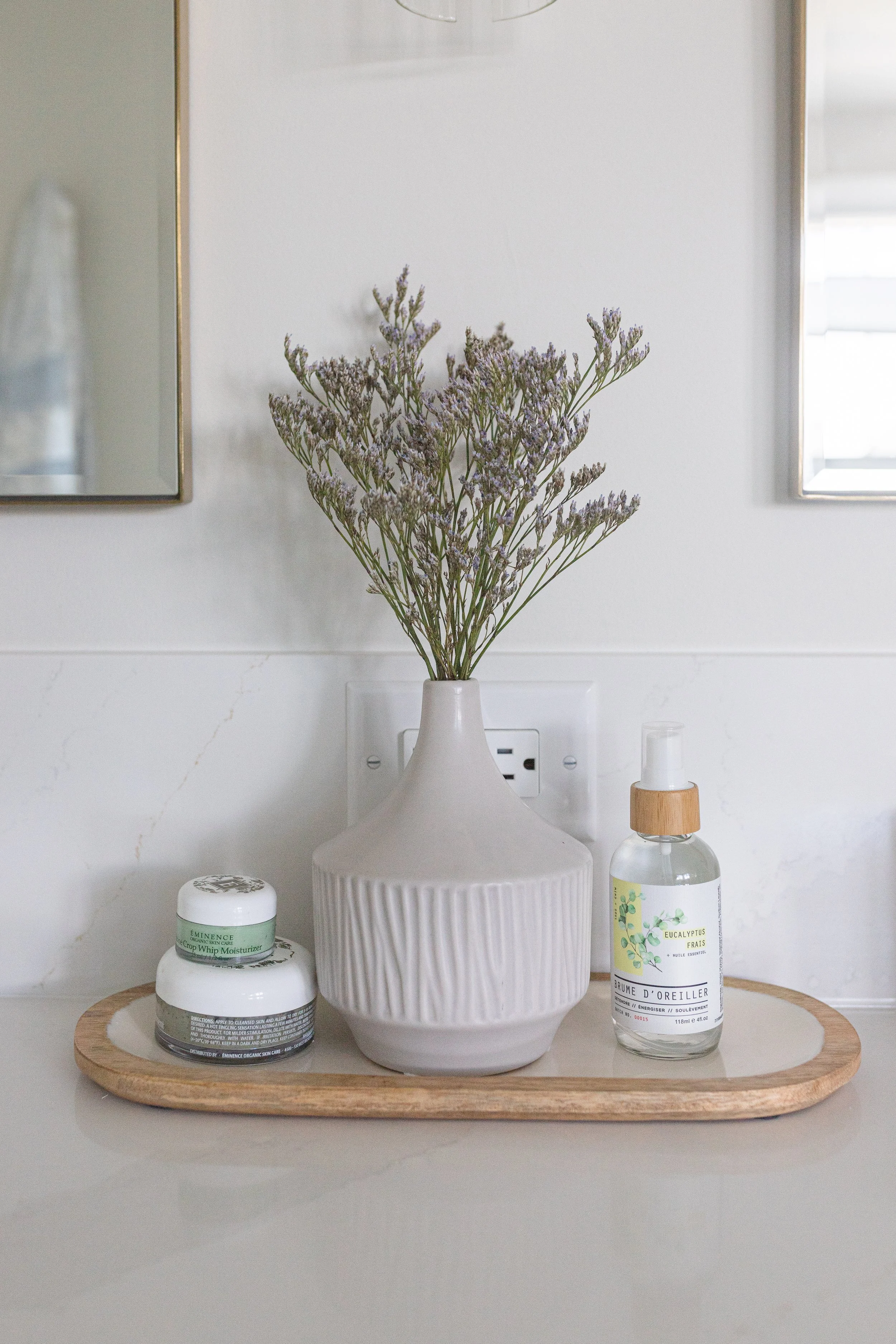 Decorative beige vase with lavender flowers, skincare products, and a spray bottle on a wooden tray, against a white wall with mirrors.