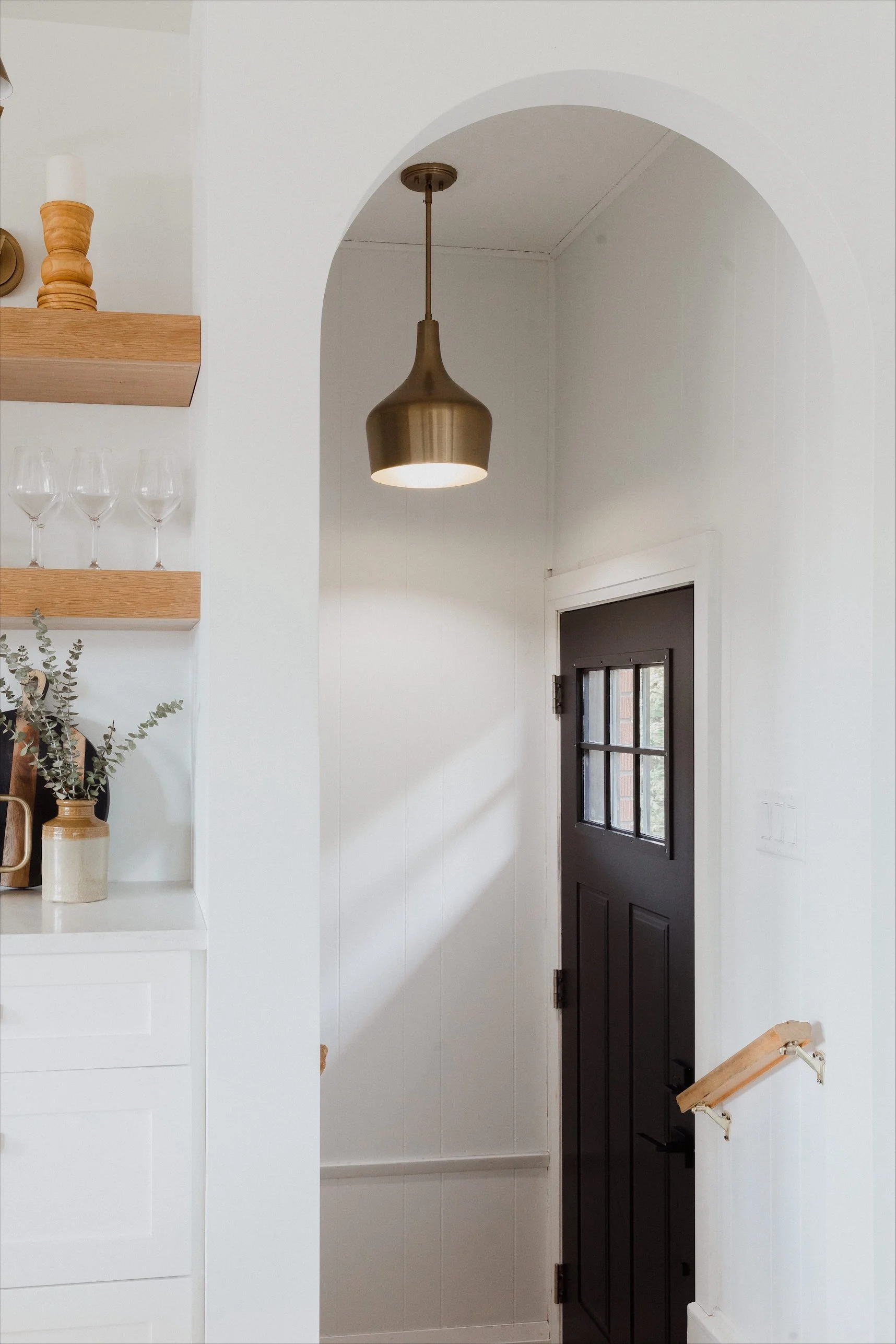 Bright bathroom with a white bathtub, glass-enclosed shower with a rain showerhead, and a window with white blinds. Decor includes a green plant, slippers, towels, a basket, and a wooden stool.