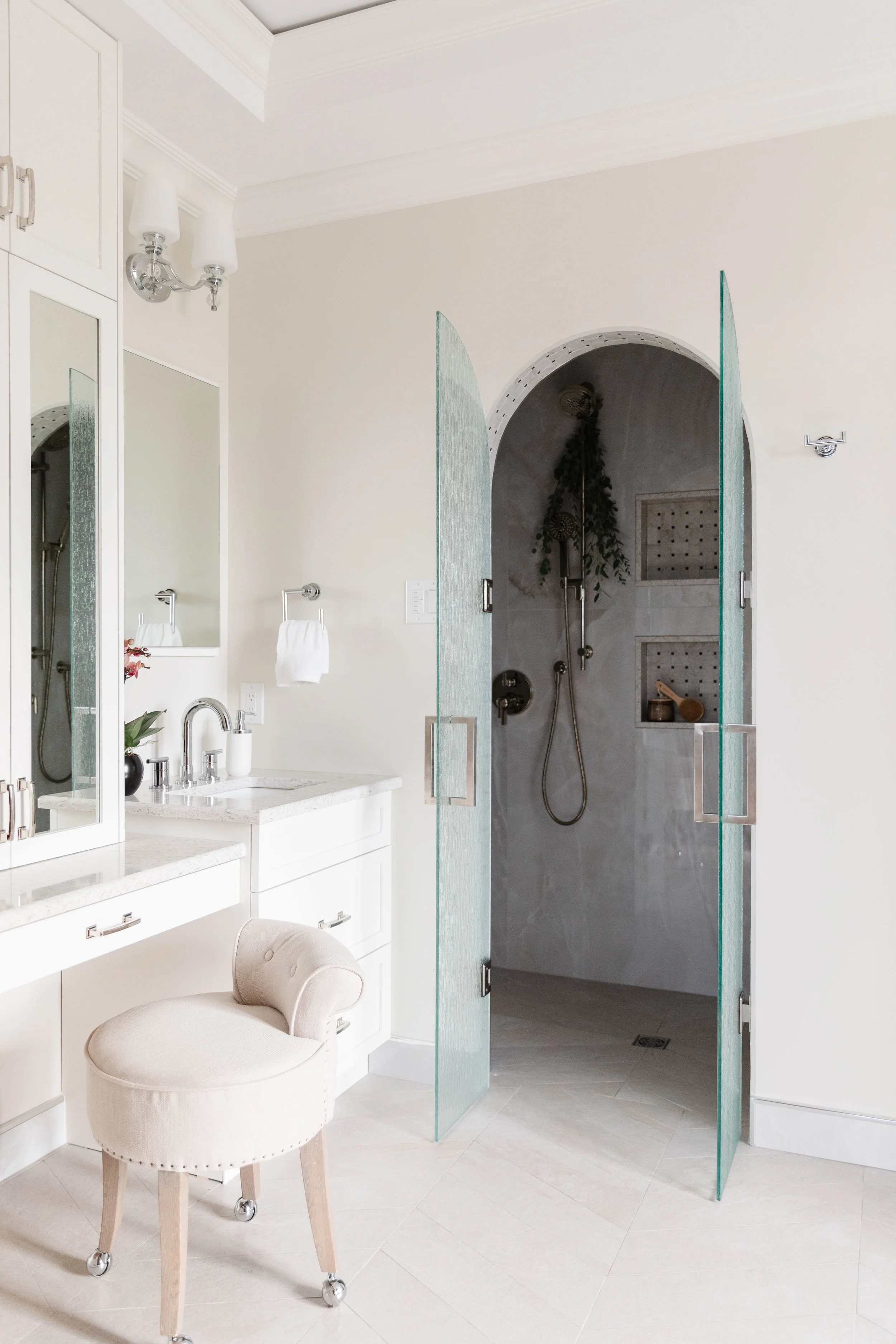 Bathroom with a shower area enclosed with glass doors, a vanity with a mirror, and a plush chair in front of the vanity.