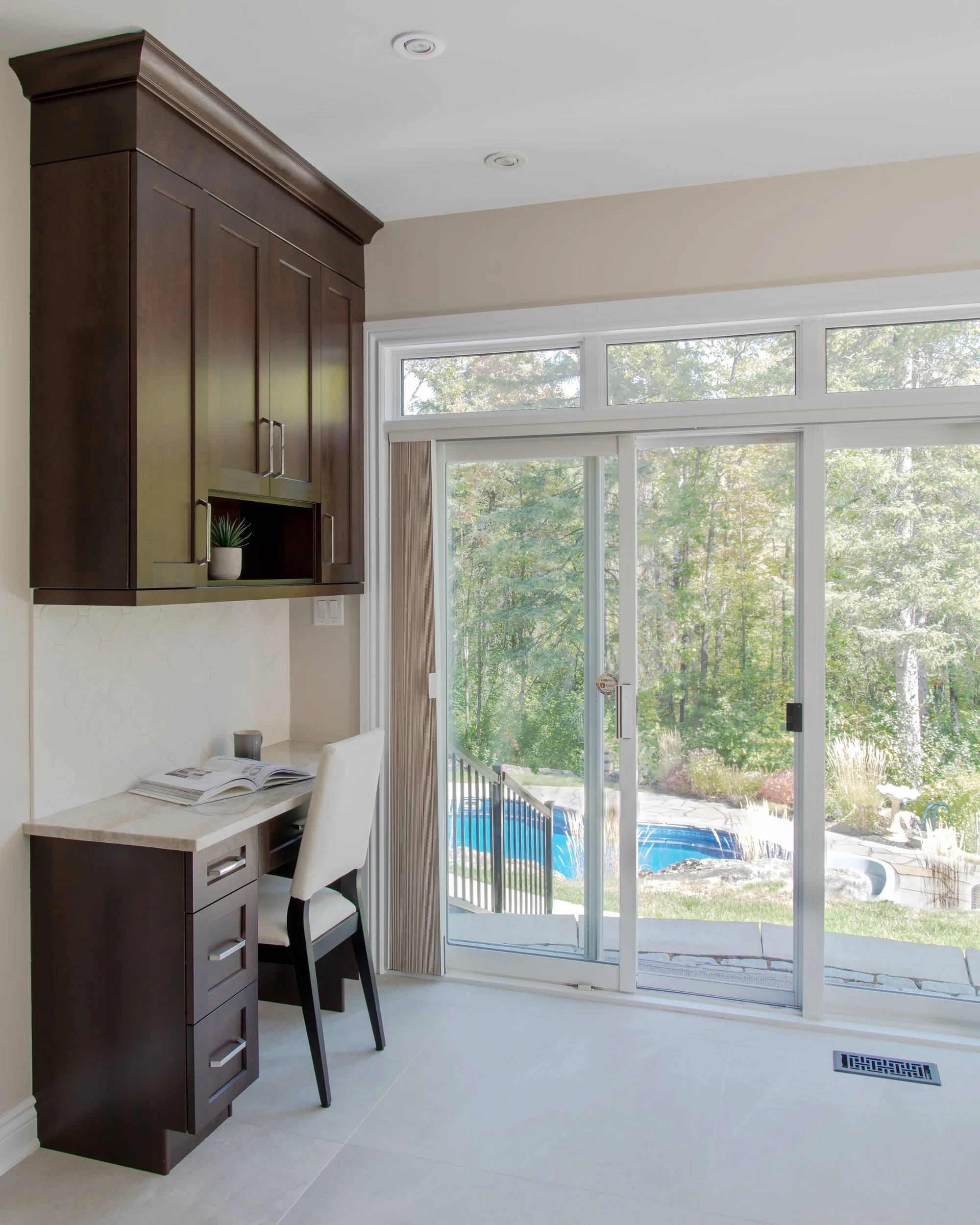 Home office area with desk, chair, and built-in dark wood cabinets facing large sliding glass door leading to backyard with pool and trees.