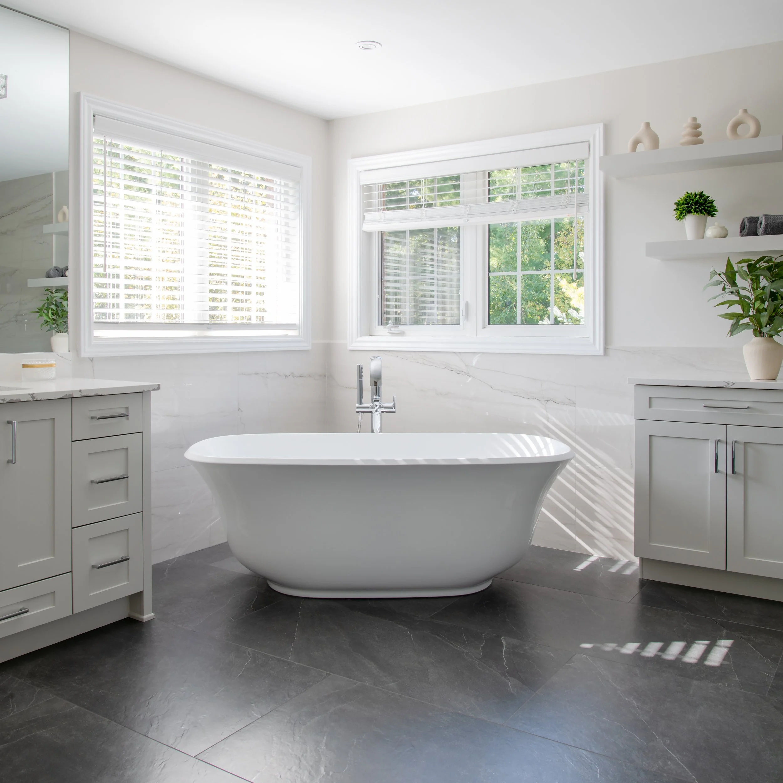 Modern bathroom with a freestanding bathtub, white cabinetry, potted plants, and large windows with blinds allowing natural light.