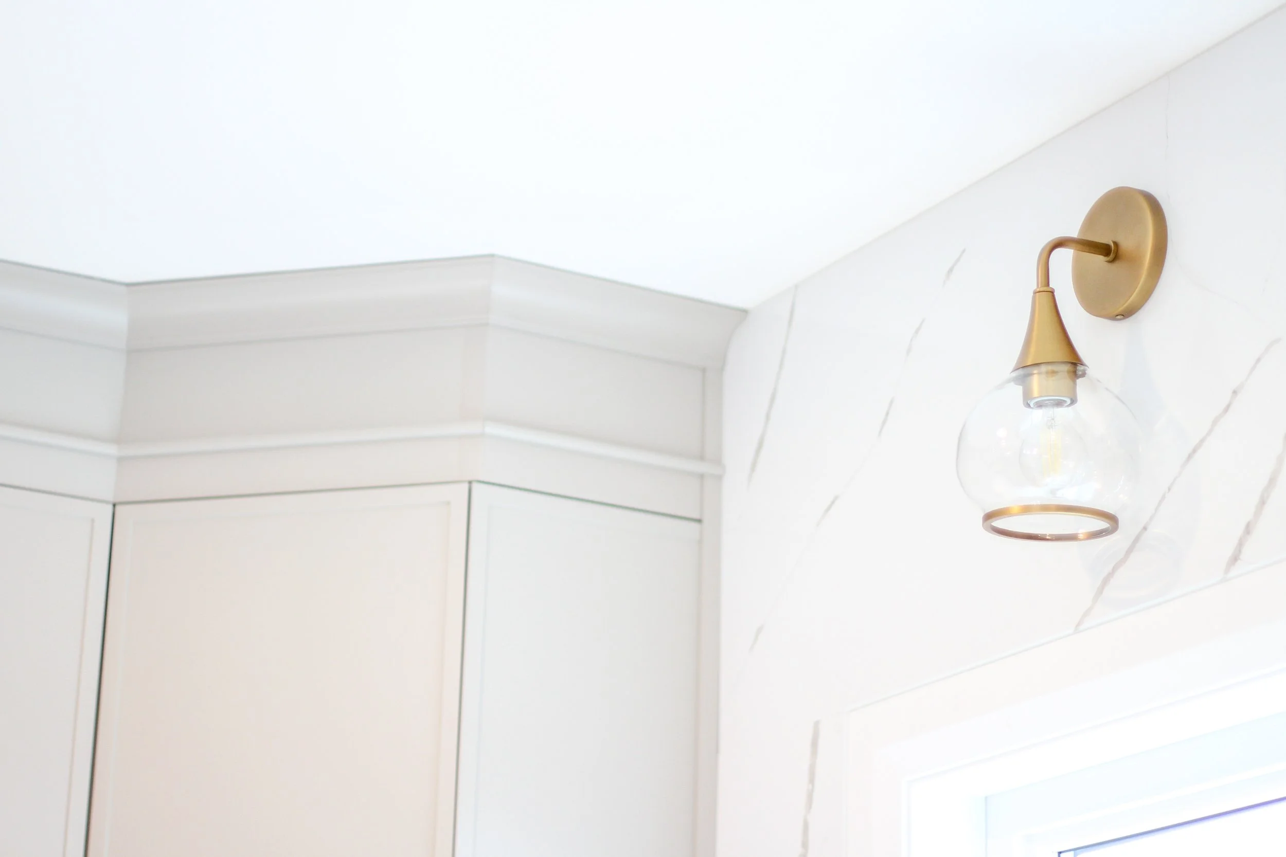 Close-up view of a modern gold and glass wall sconce mounted on a white wall with subtle marble pattern, near a corner with a ceiling and window.