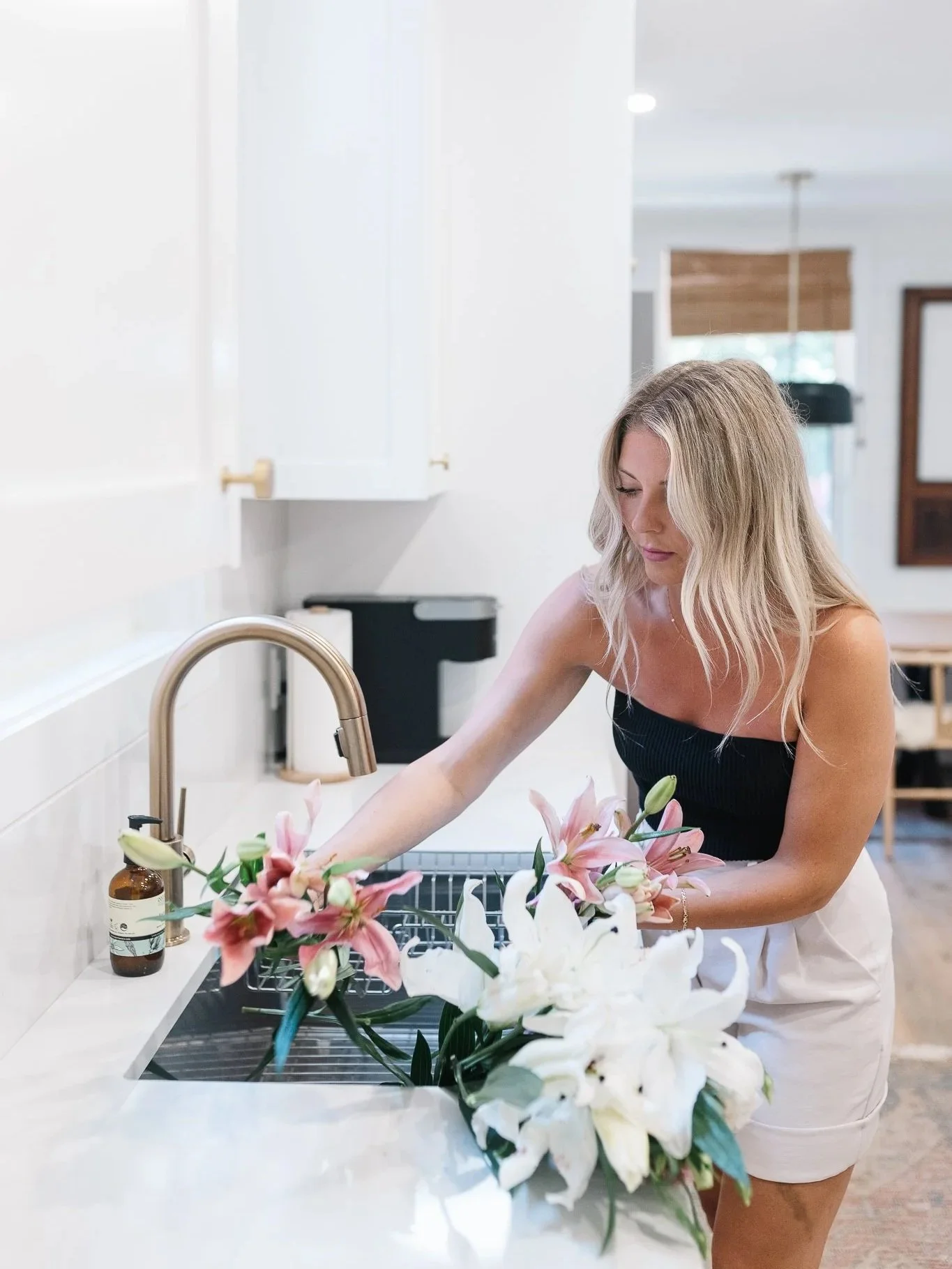 Woman with blonde hair arranging a bouquet of pink and white lilies in a kitchen sink.