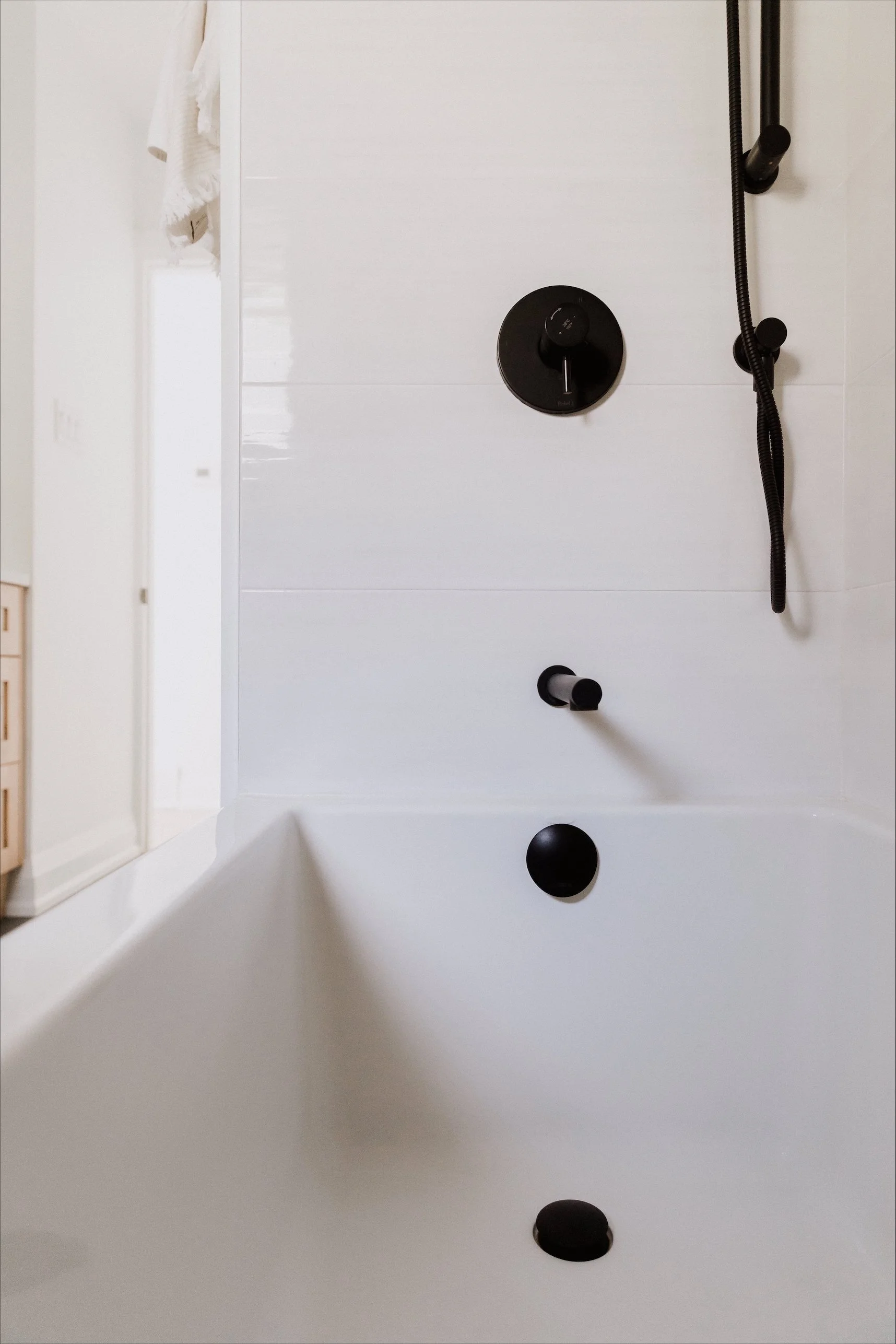 Modern bathroom with light-colored walls and flooring, featuring a large mirror above a white vanity with gold hardware, a bathtub with a glass partition, and a ladder towel rack near a window, decorated with minimalistic art and decor.