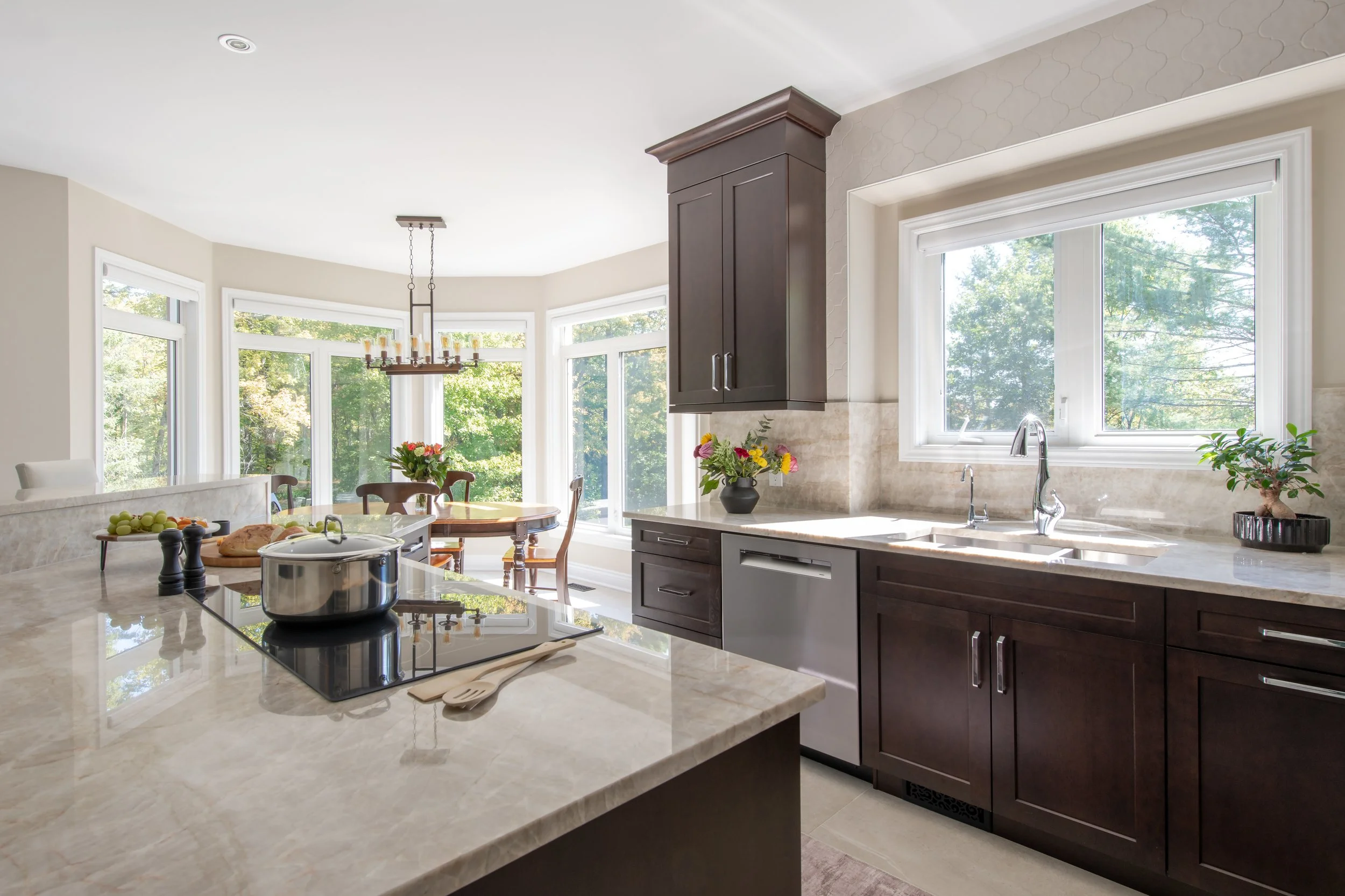 Bright kitchen with dark wood cabinets, marble countertops, a window above the sink, and a dining area with a round table and chairs in a sunny, green outdoor view.