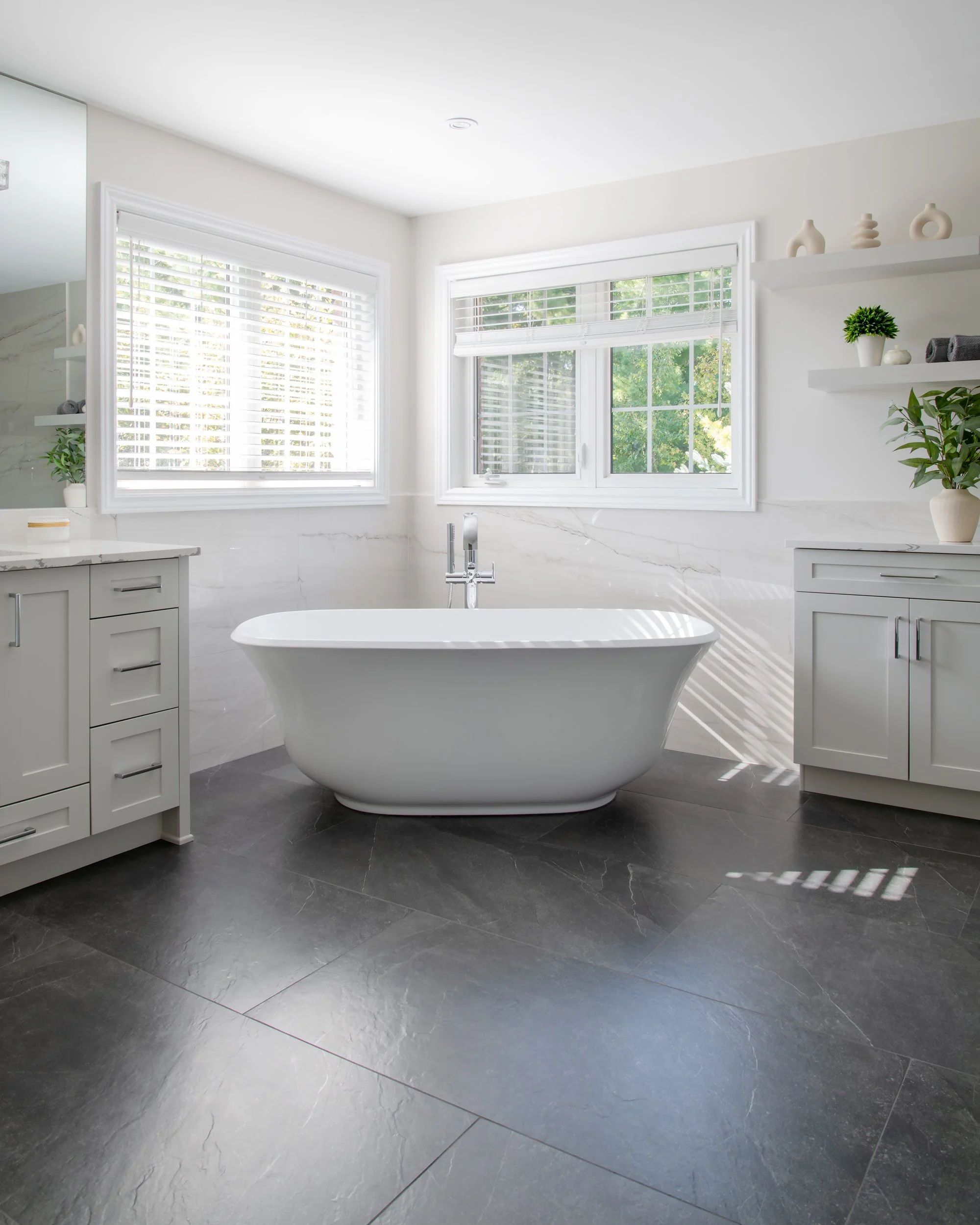 Modern bathroom with white freestanding bathtub, two large windows with white blinds, gray cabinets, and decorative plants on shelves