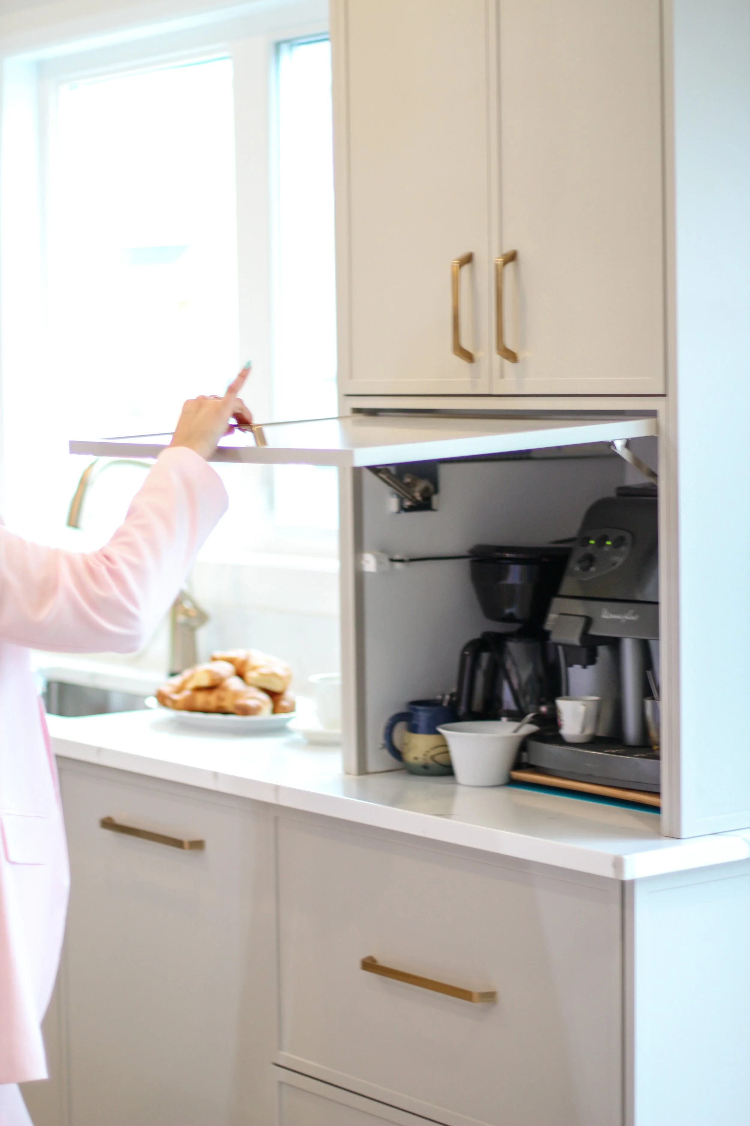 Person opening a kitchen cabinet above a white countertop with coffee maker, bowls, and mugs, with a plate of pastries nearby, in a bright kitchen with windows.