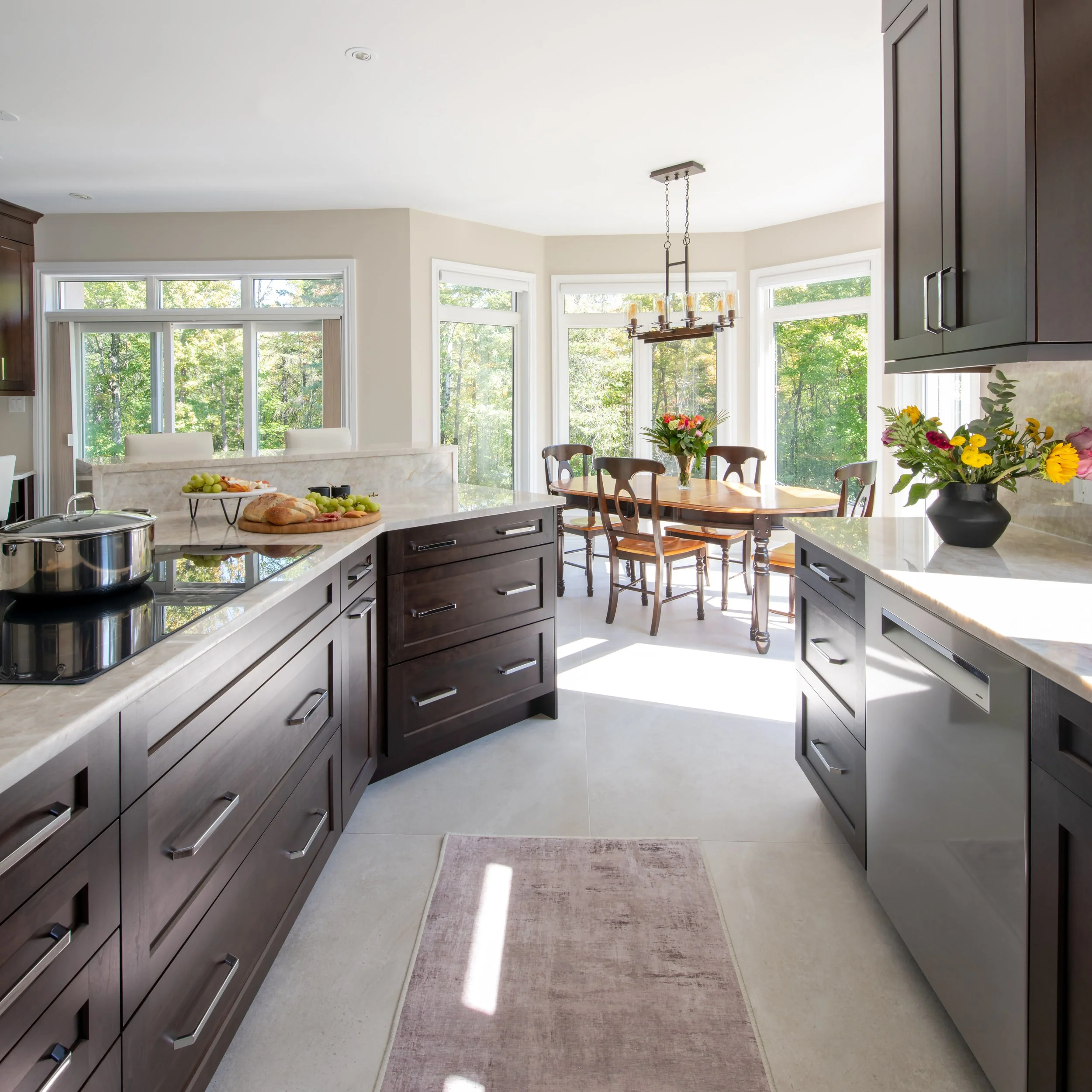 Bright and airy kitchen with dark wood cabinets, light countertops, and a view of a sunny dining area with a wooden table, chairs, and a chandelier surrounded by large windows with trees outside.